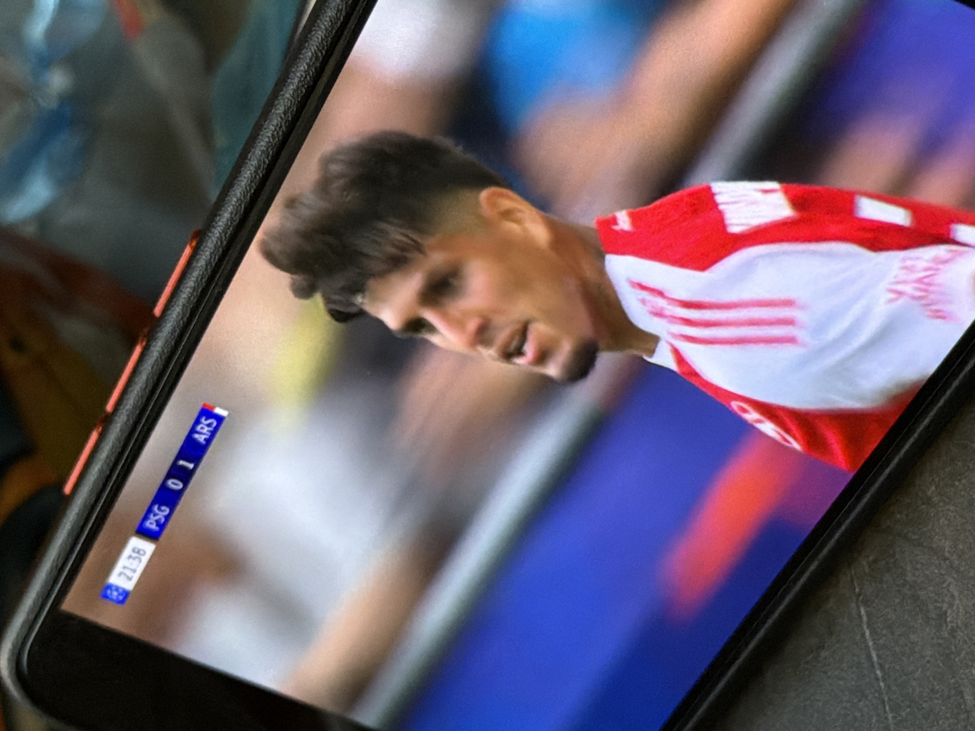 The image shows a smartphone displaying a soccer game between PSG and ARS (Arsenal) with the score being 0-1 in favor of Arsenal at the 71:38 mark. The screen shows a player in a red and white jersey. The phone is lying on a flat, gray surface. There is no evidence to confirm the image was taken in the city of Chaikovsky, Russia.