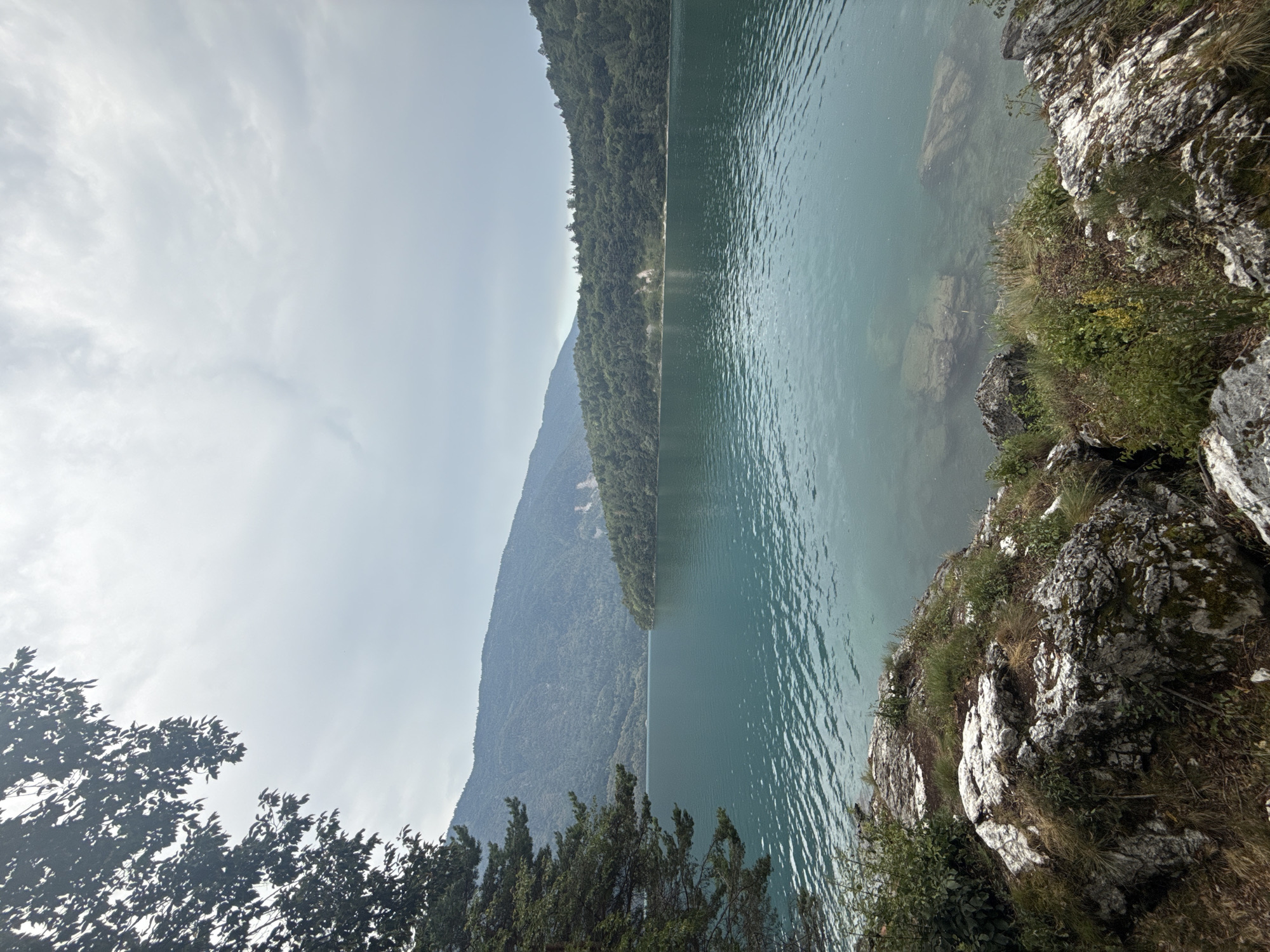 An outdoor, daytime, vertical, eye-level shot captures a serene lake with tranquil, turquoise water and a distant mountainous landscape under a cloudy sky. The foreground features a rocky shore with sparse green vegetation. The midground is dominated by the expansive lake, reflecting the muted light of the overcast sky. In the background, lush green forests cover rolling hills that rise to meet the hazy, gray mountains. The scene is devoid of any human presence, artificial structures, or vehicles, creating a natural and peaceful ambiance. The overall impression is one of untouched natural beauty.