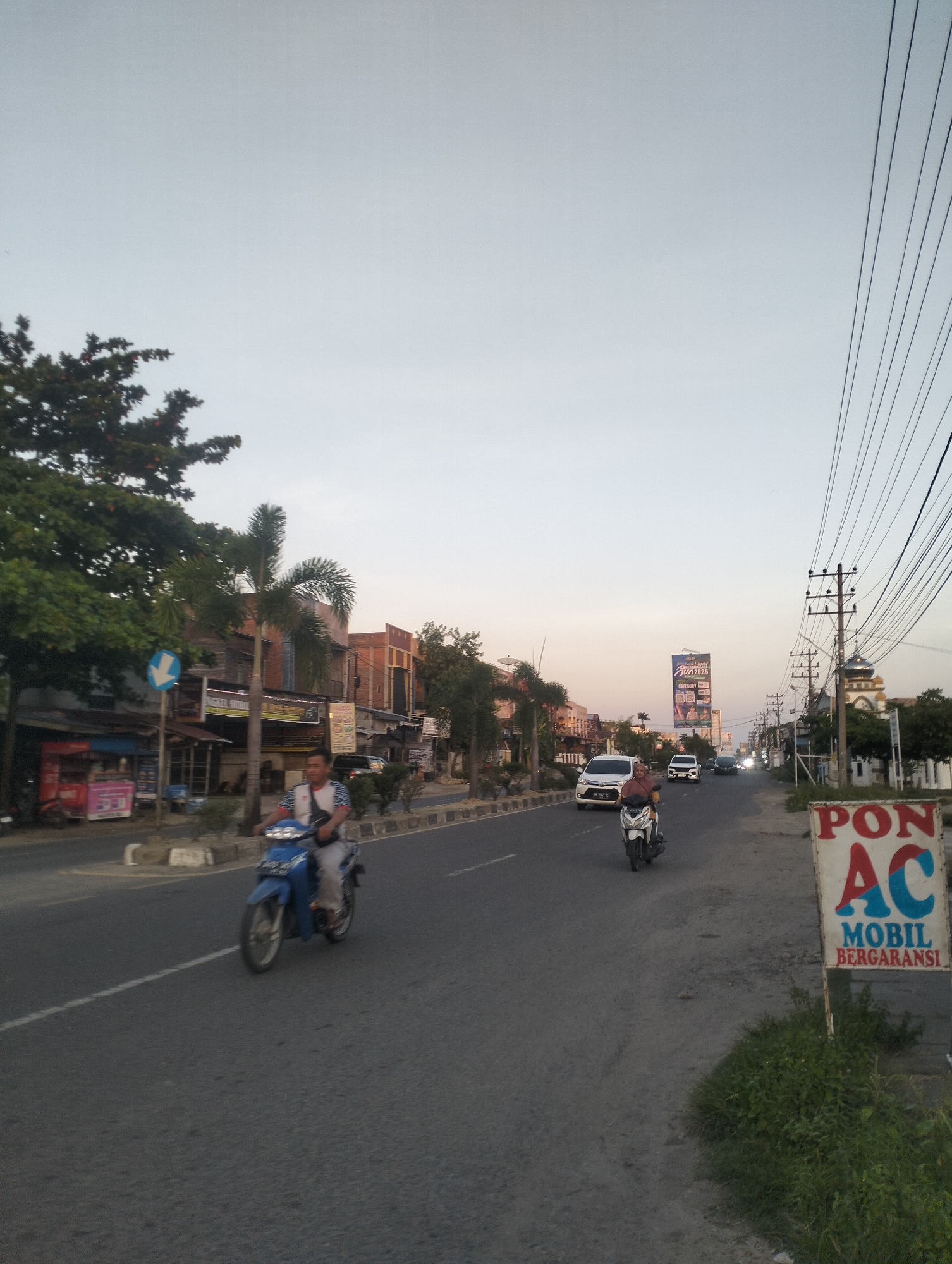 This is a horizontal, eye-level shot of a street in Lhokseumawe, Indonesia. The sky is overcast and light blue. On the right side of the road, a billboard reads "PON AC MOBIL BERGARANSI." The road is paved and has a white line down the center. There are several vehicles on the road, including a blue motorcycle with a person on it in the foreground, a white car, and several other vehicles in the distance. On the left side of the road are several buildings, including shops and what appears to be a mosque with a dome. Trees and power lines are also visible. The overall impression is one of a busy street scene in a tropical urban environment.