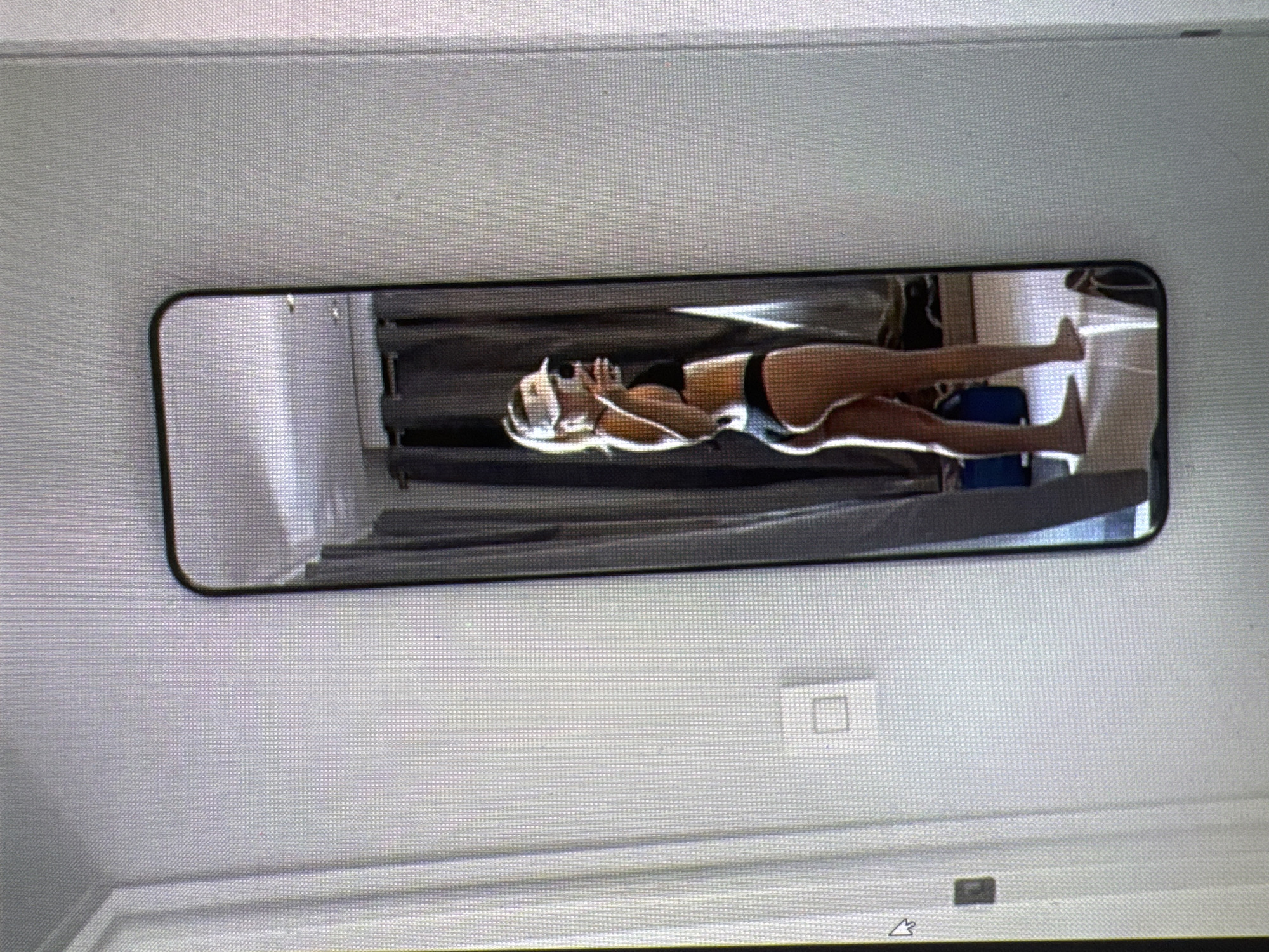 The media file is a photograph of a full-length mirror reflecting a person. The person appears to be a woman lying down, wearing a black bikini. She is positioned horizontally within the mirror's frame. The background visible in the reflection suggests an indoor setting, possibly a bedroom or dressing area, with what looks like a curtain or partition.
The image is captured at an angle, with the mirror itself having a black frame. The overall lighting appears to be artificial, and no specific time of day or weather conditions can be determined. The reflection shows the person's legs and torso, with her hands near her face. There are no discernible emotional cues from the person's pose.
The photograph is taken from a slight distance, with the mirror occupying a significant portion of the frame. The wall surrounding the mirror is a light gray color and textured. There are no visible signs or text in the image itself. The location context provided is Castelo Branco, Portugal, but there are no visual cues within the image to confirm this.
