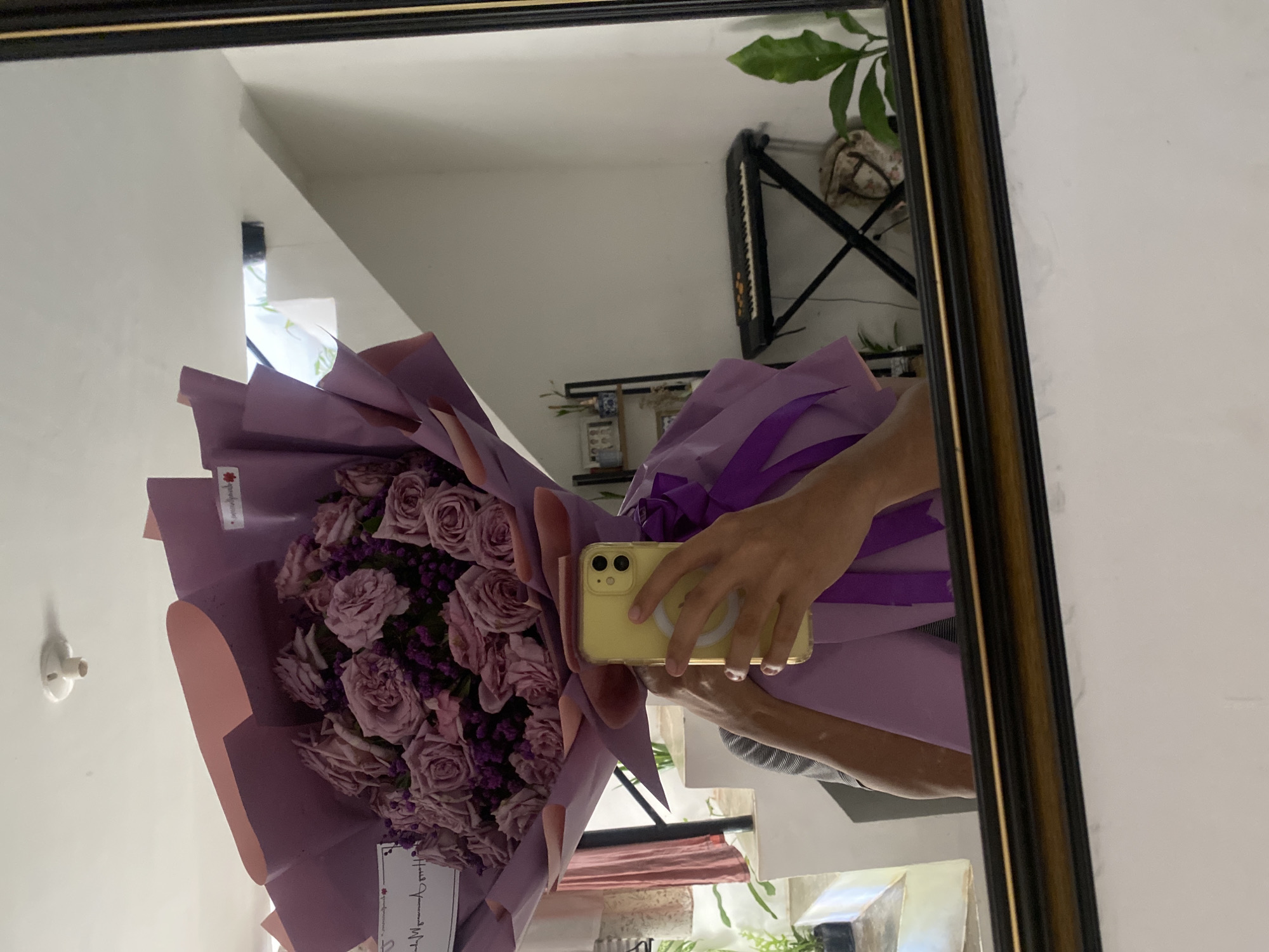 The scene is a reflection in a mirror of a person holding a bouquet of purple roses and small purple flowers wrapped in purple paper. The person is holding a yellow iPhone with a visible MagSafe circle on the back. The reflection reveals part of an interior space, including a white wall, a plant on a black metal stand, and a keyboard or synthesizer. The lighting in the room appears bright. The reflection suggests an indoor location. There is no evidence of the city of Davao City, Philippines.