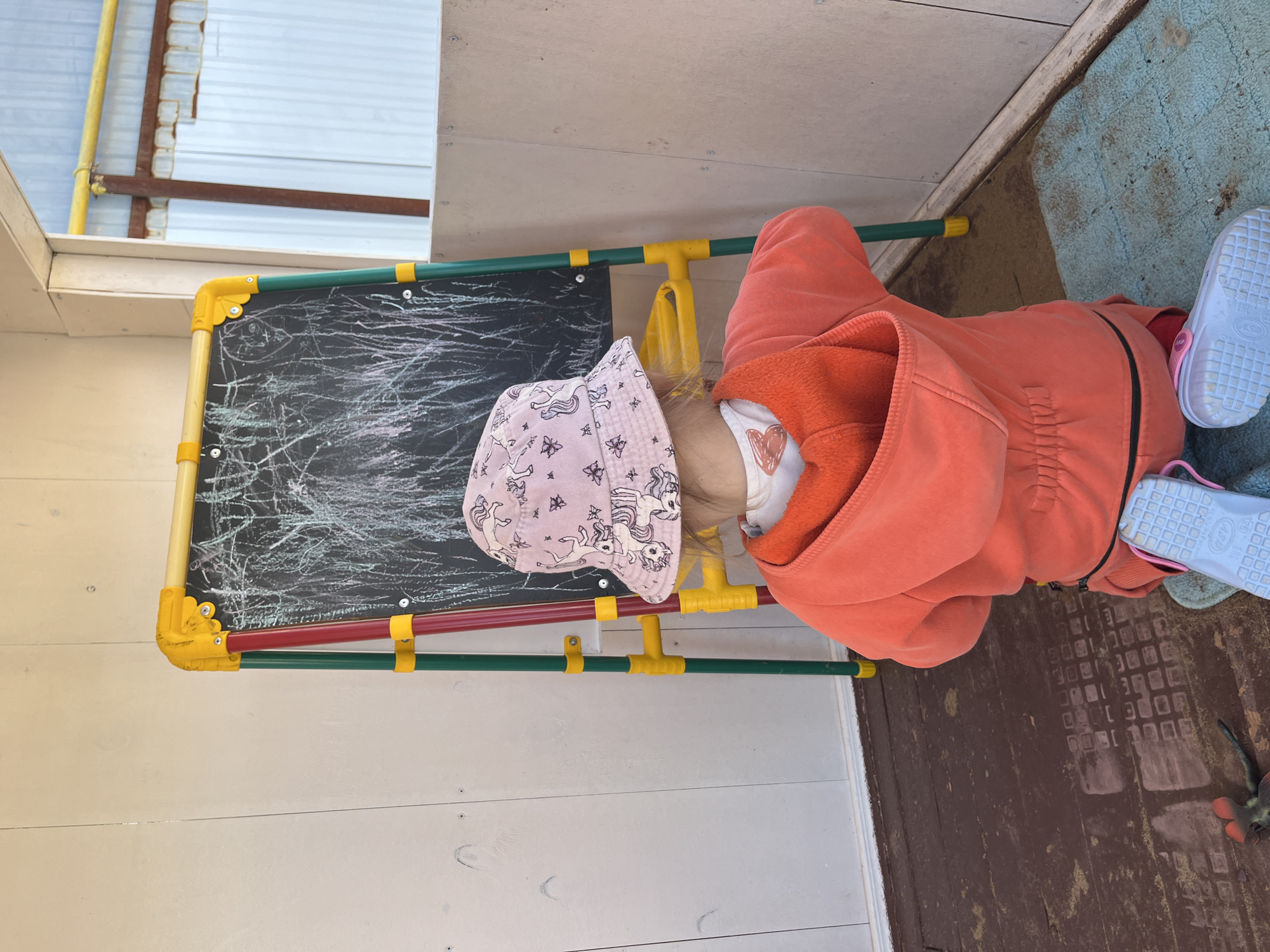 The image depicts a young child from behind, kneeling on a light blue mat and engaged with a colorful chalkboard easel, in what appears to be an enclosed or semi-enclosed outdoor space, such as a veranda or porch, in the city of Chaikovsky, Russia.
The child wears a pink bucket hat patterned with unicorns and butterflies, an orange hooded jacket with a darker orange interior lining, and red pants. Light blue plastic clogs with pink straps are on the child's feet, resting on the mat. Light-colored hair is visible beneath the hat. The child's posture indicates drawing or interacting with the chalkboard.
The chalkboard easel, positioned directly in front of the child, presents a black drawing surface extensively covered with white and light-colored chalk marks, comprising scribbles, vertical lines, and abstract shapes, consistent with a child's artwork. The easel's frame is constructed from yellow, red, and green plastic or painted metal components, with yellow connectors linking the red and green support legs.
The immediate environment includes light-colored, vertically paneled wooden walls on the left and behind the easel. The wall on the right is a lighter, possibly unfinished wooden panel. Visible screws are present in the wall panels. The floor consists of dark brown wooden planks, showing signs of wear and scuff marks. To the lower left, distinct rectangular patterns, likely from shoe soles, are imprinted into the dusty floor. A small, dark object with red accents, possibly a toy, rests on the wooden floor to the child's left. Beyond an opening on the right, an exterior scene includes corrugated metal paneling, a horizontal yellow pipe positioned high, and a vertical rusty brown beam, indicating an adjacent structure or fence.
