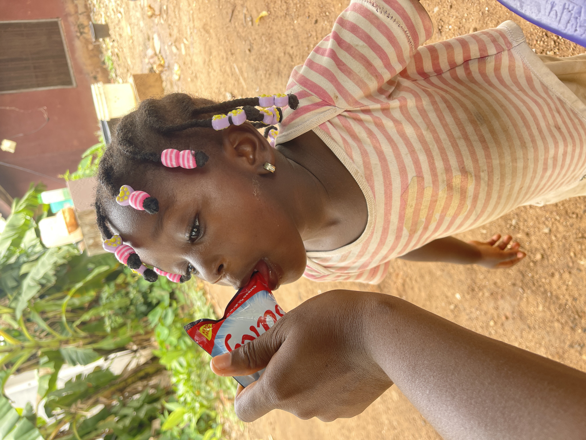 The image presents a close-up, slightly tilted shot of a young Black girl consuming a product from a sachet held by an adult's dark-skinned hand. The scene is set outdoors in Akwanga, Nigeria, under bright daylight.
The main subject is the young girl, seen from her chest up. She has dark, braided hair adorned with numerous colorful beads, including pink and white striped bow-like designs and light purple and yellow beads. She wears a light-colored top with horizontal pink and white stripes, which exhibits some faint smudges or stains. A small, gold-colored hoop earring is visible in her left ear. Her eyes are open and gaze slightly upwards as she brings her mouth to the packet, her tongue visible as she extracts the contents.
An adult's hand, with dark skin, holds a small, flexible sachet for the girl. The packet is predominantly red, white, and blue. Visible text on the sachet includes "NEW PACK" and a partial phrase "- % GERMS," along with what appears to be part of a brand name.
The background shows reddish-brown earth covering the ground. On the left side of the frame, vibrant green tropical foliage is visible, possibly banana leaves or similar plants. In the upper left background, a faded reddish-brown building or wall with a window can be seen. The bright, even lighting suggests it is daytime. The overall image captures a casual, everyday moment of a child enjoying a snack or drink.