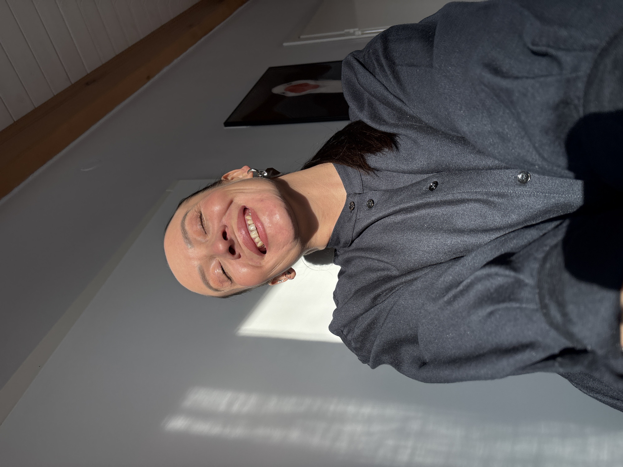 A woman with dark hair is smiling and looking directly at the camera. She is wearing a dark grey button-down shirt. The background is a white wall with sunlight shining on it, and an angled ceiling with a dark painting hanging on the wall. The location appears to be indoors. There is no specific information to indicate that the picture was taken in the city of Chaikovsky, Russia.