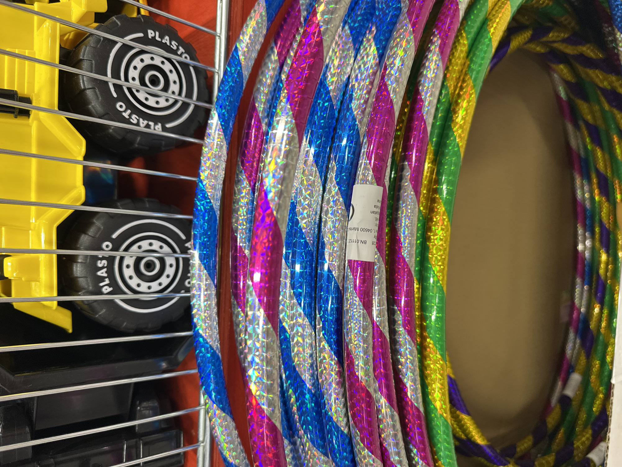 A close-up, eye-level shot shows a stack of colorful hula hoops in a wire basket. The hula hoops are covered in iridescent foil that reflects light, creating a shimmering effect. They are striped with alternating colors, including blue, pink, silver, green, yellow, and purple. In the background, partially obscured by the basket and hoops, is a yellow toy truck with black wheels that have "PLASTO" printed on them. The truck appears to be a large, commercial toy, possibly in a store display. The lighting is bright, highlighting the reflective surfaces of the hoops. The overall scene is vibrant and playful, suggesting a retail environment or a toy display.