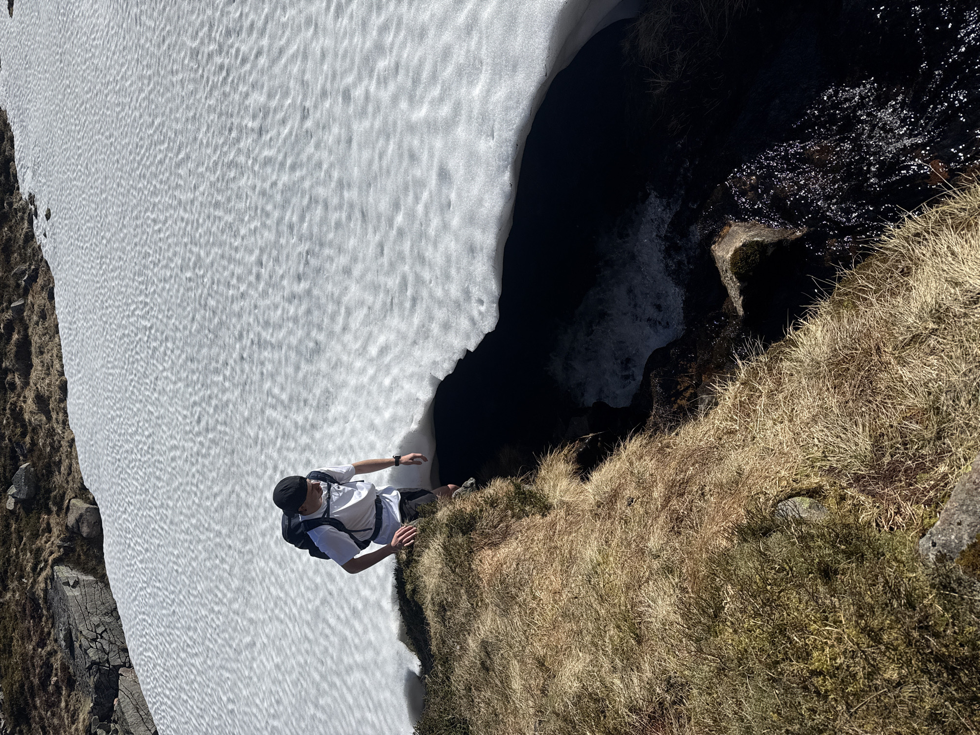 A man in a white t-shirt, black shorts, a black cap, and a backpack stands on the edge of a grassy bank. To his left is a snowfield, and to his right, there is a dark body of water, possibly a stream or a river. The location is unknown.