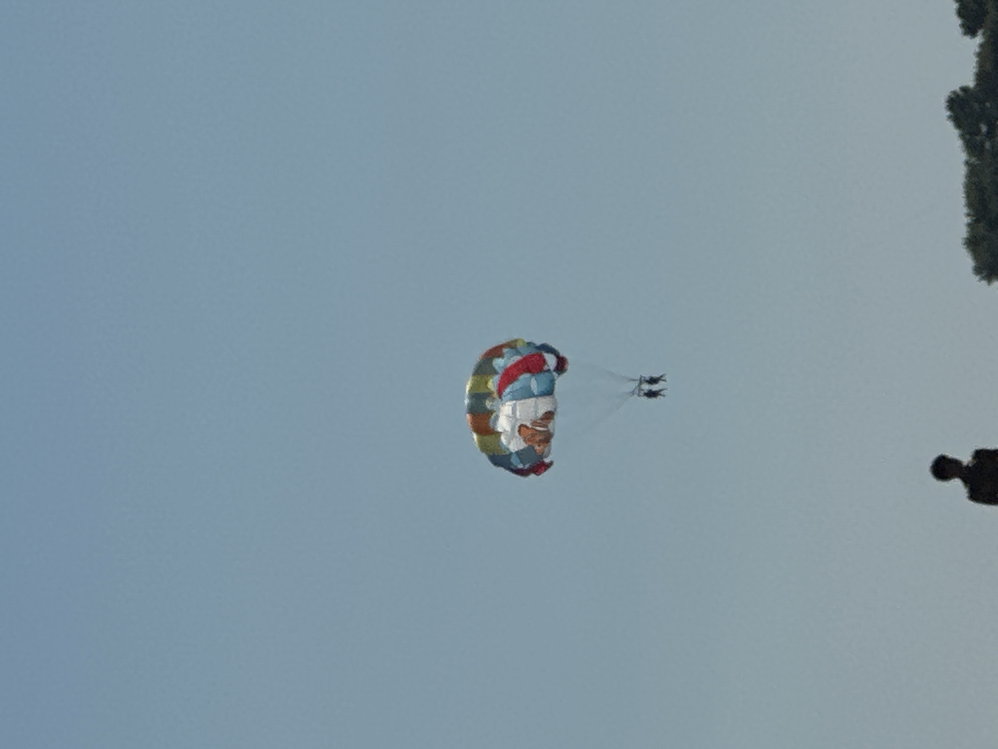 The image shows a clear, light blue sky with a parachute in the upper center. The parachute is colorful, with segments of red, white, blue, yellow, and green. Attached to the parachute are two individuals, appearing as dark silhouettes against the sky. They are suspended in the air, presumably parasailing. In the foreground, near the bottom of the image, the silhouette of a person is visible. Below this silhouette, at the very bottom edge of the frame, a dark green, leafy tree line is present, suggesting this scene is taking place outdoors, possibly near a coast or body of water where parasailing activities are common. The overall environment appears to be a bright, clear day. The location in Boreti, Montenegro, implies this is likely a coastal or lakeside area.