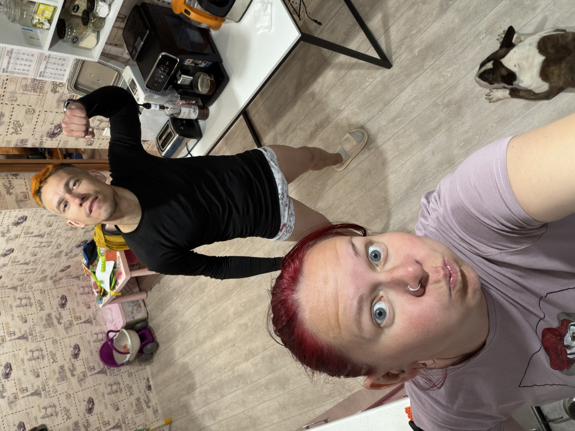 The image appears to be a selfie taken indoors, likely in a kitchen, as indicated by the presence of kitchen appliances and cabinets in the background. Two individuals are visible: one male and one female. The male has orange hair, is wearing a long-sleeved black shirt, and is flexing his bicep. The female has red hair and is wearing a light purple shirt with a cartoon image on it; she is making a surprised facial expression. A white and brown dog is lying on the floor. Other objects in the background include a white table, a toaster, a microwave, and cabinets filled with jars. A child's play table and toy storage are also visible.
The location is in the city of Chaikovsky, Russia.