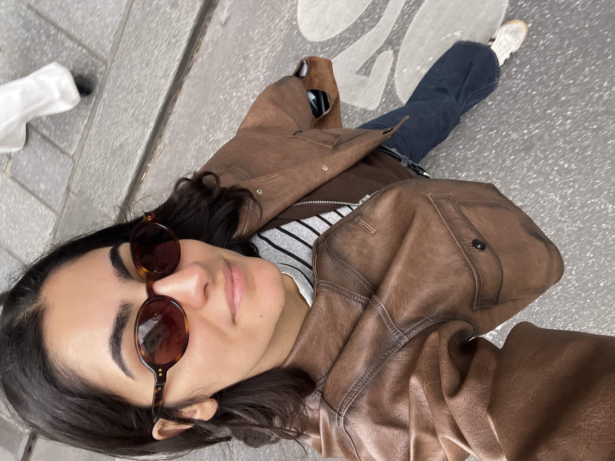 A medium-angle, high-perspective selfie captures a young adult female from the chest down, positioned on a grey paved street in the Montorgueil district of Paris, France.
The subject has dark, wavy hair and wears oval-framed, tortoise-shell sunglasses with dark lenses; urban architecture is reflected in the lenses. She is clad in a brown, textured leather jacket with prominent stitching, visible zippers, and a buttoned flap pocket on the wearer's right side. Beneath the jacket, a grey and white horizontally striped shirt is visible at the collar, layered over a solid brown top. She wears dark blue jeans and white sneakers with light-colored soles and a subtle red detail on the heel of the right shoe. A dark belt buckle is visible above the jeans. A small, open brown bag or pouch is tucked under her right arm, revealing dark objects inside.
The ground is composed of large, light grey rectangular paving stones. Distinctive white, abstract, and flowing line patterns are painted on the street surface, characteristic of pedestrian zones such as Rue Montorgueil in Paris. In the upper right background, the lower legs and feet of another person are visible, wearing loose white trousers and dark shoes, walking away from the camera on the same paved surface.