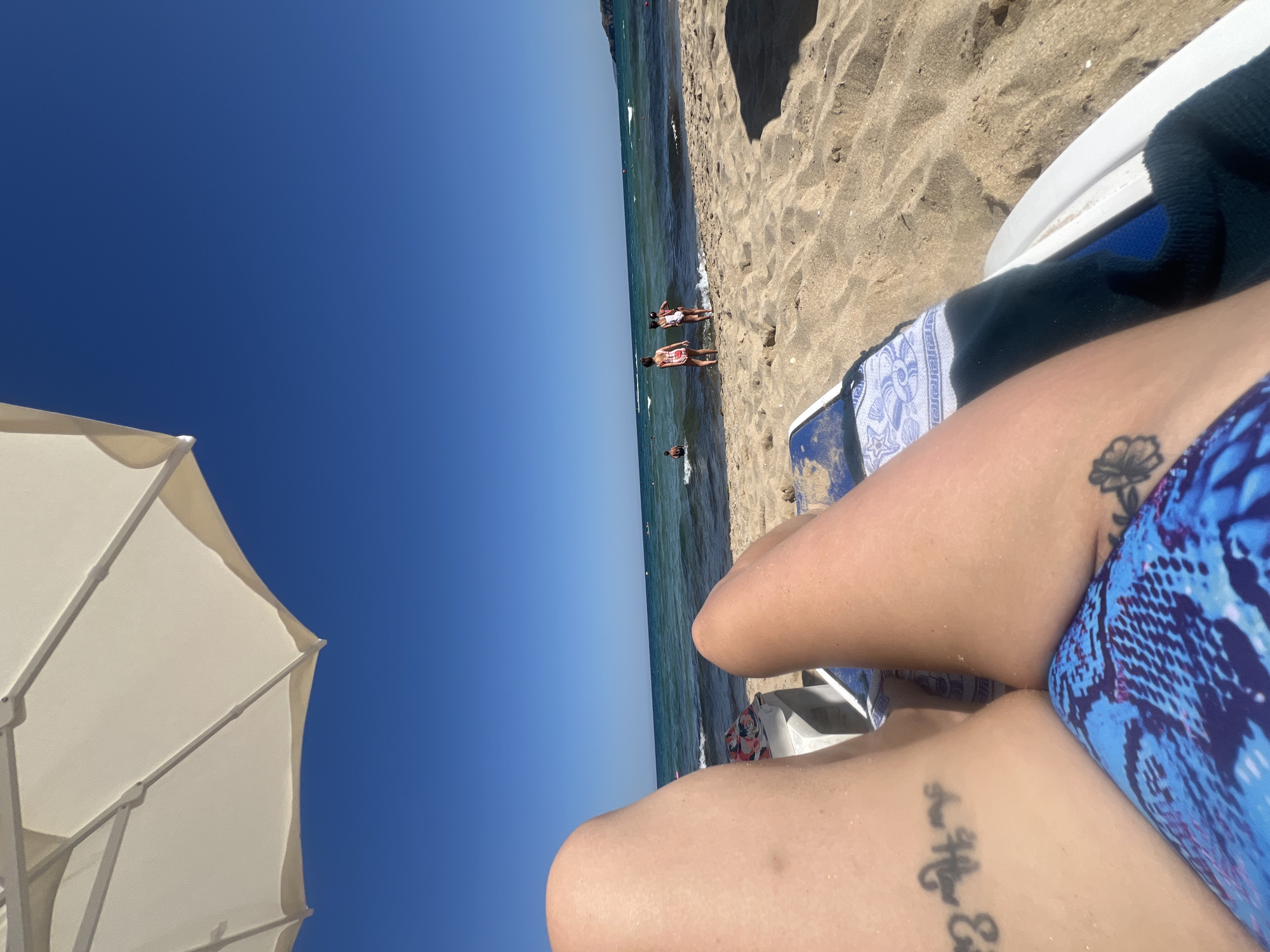 The image depicts a first-person perspective of someone relaxing on a beach chair on a sandy beach. The foreground is dominated by the legs of the individual, showing tattoos on the left thigh and right calf. They are wearing blue patterned swimwear. A white beach umbrella is visible in the upper left corner, providing shade. The midground features the ocean with gentle waves and several people in the water, including children. The background shows a clear blue sky and a distant coastline. The setting is a beach in Sozopol, Bulgaria.