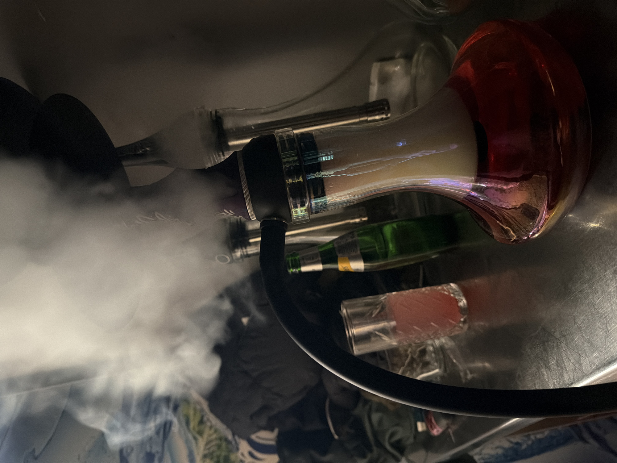 The image depicts a lit hookah on a reflective surface surrounded by various objects, including glasses and bottles. Smoke is emanating from the hookah hose. The hookah has a red and clear glass base with a black stem and hose. The surrounding environment appears cluttered, with miscellaneous items scattered around the hookah. There is no information in the image that directly identifies the location as Varna, Bulgaria.