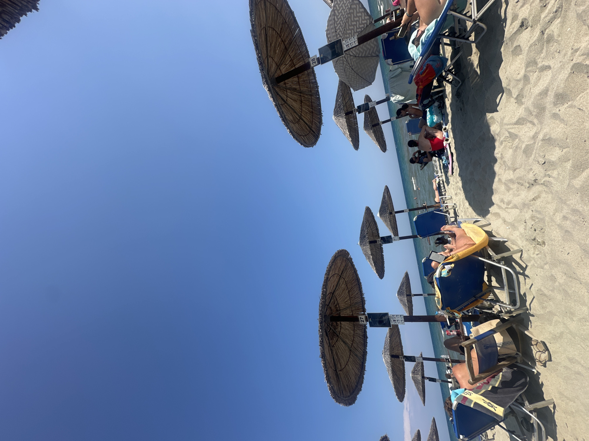 The image depicts a sunny day at a beach in an unknown location in Greece. The foreground is dominated by sandy beach with several beach chairs and umbrellas. In the midground, people are seen relaxing on beach chairs, some reading, others chatting. The ocean is visible in the background with gentle waves. Several straw umbrellas are positioned on the sand, providing shade. Some umbrellas have numbers visible on their posts, such as '27' and '28'. The sky is clear and blue.