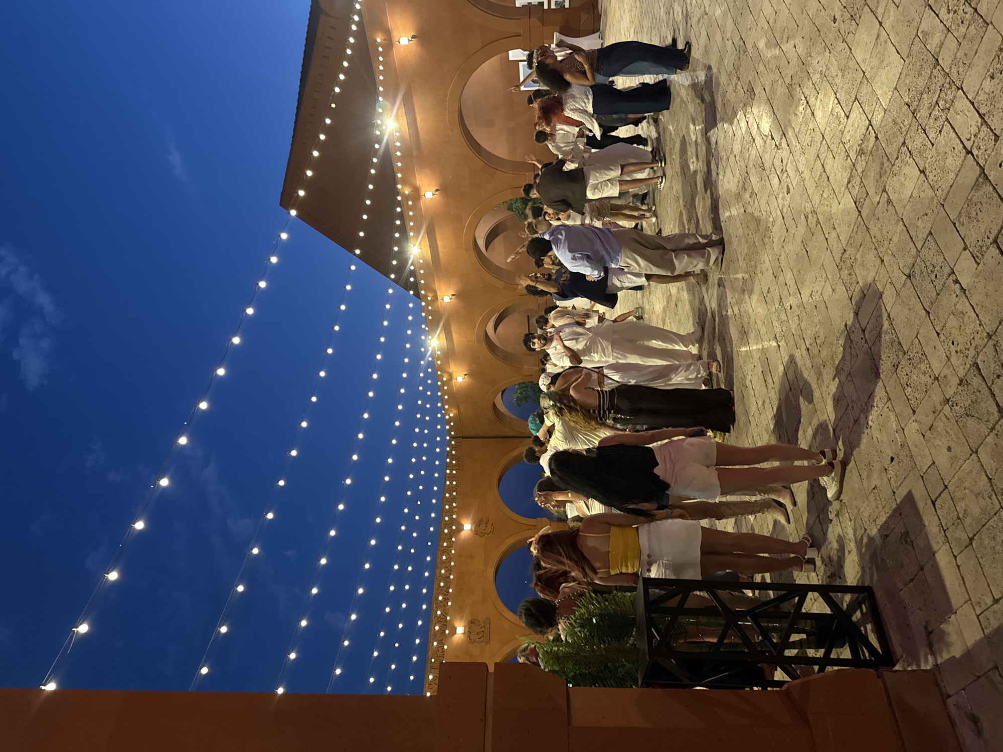 The image depicts an outdoor evening gathering at a venue in Sasunik, Armenia, illuminated by strings of lights. A crowd of people, dressed in casual summer attire, are socializing and dancing on a paved courtyard. The background features a large, terracotta-colored building with arched openings and a distinctive roofline. The sky above is a deep blue, indicating it is dusk. The atmosphere appears festive and relaxed, with individuals engaging in conversations and enjoying the event.