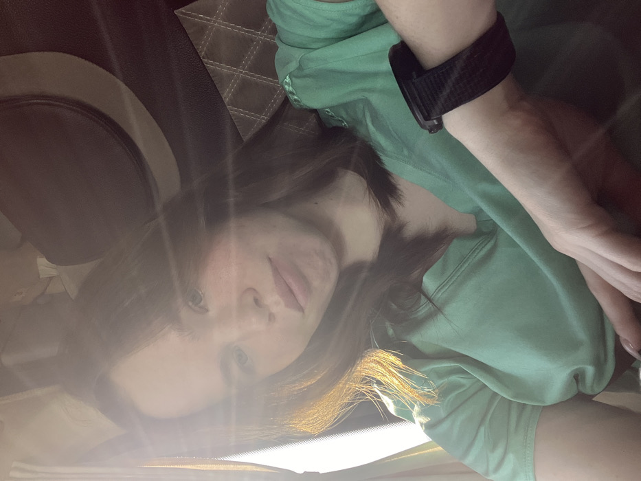 The image depicts a single female subject within what appears to be the interior of a vehicle, stated to be located in Lipovka, Russia. The subject, an adult female with long, dark brown hair, is positioned facing forward, slightly angled towards the camera. Strong natural light, originating from the left side of the frame, casts a bright flare across the upper left quadrant of the image and illuminates the left side of her face and hair. She wears a light green short-sleeved top with a V-neck. A black digital wristwatch with a black textured fabric strap is visible on her right wrist. Her right hand is partially visible in the lower right, resting in front of her. The visible background consists of an upholstered headrest and seatback, both dark-colored, with the seatback featuring a diamond-quilted pattern. The overall lighting suggests a daytime setting. The subject's expression is neutral as she looks directly at the camera. No discernible action or interaction is taking place beyond posing for the photograph.