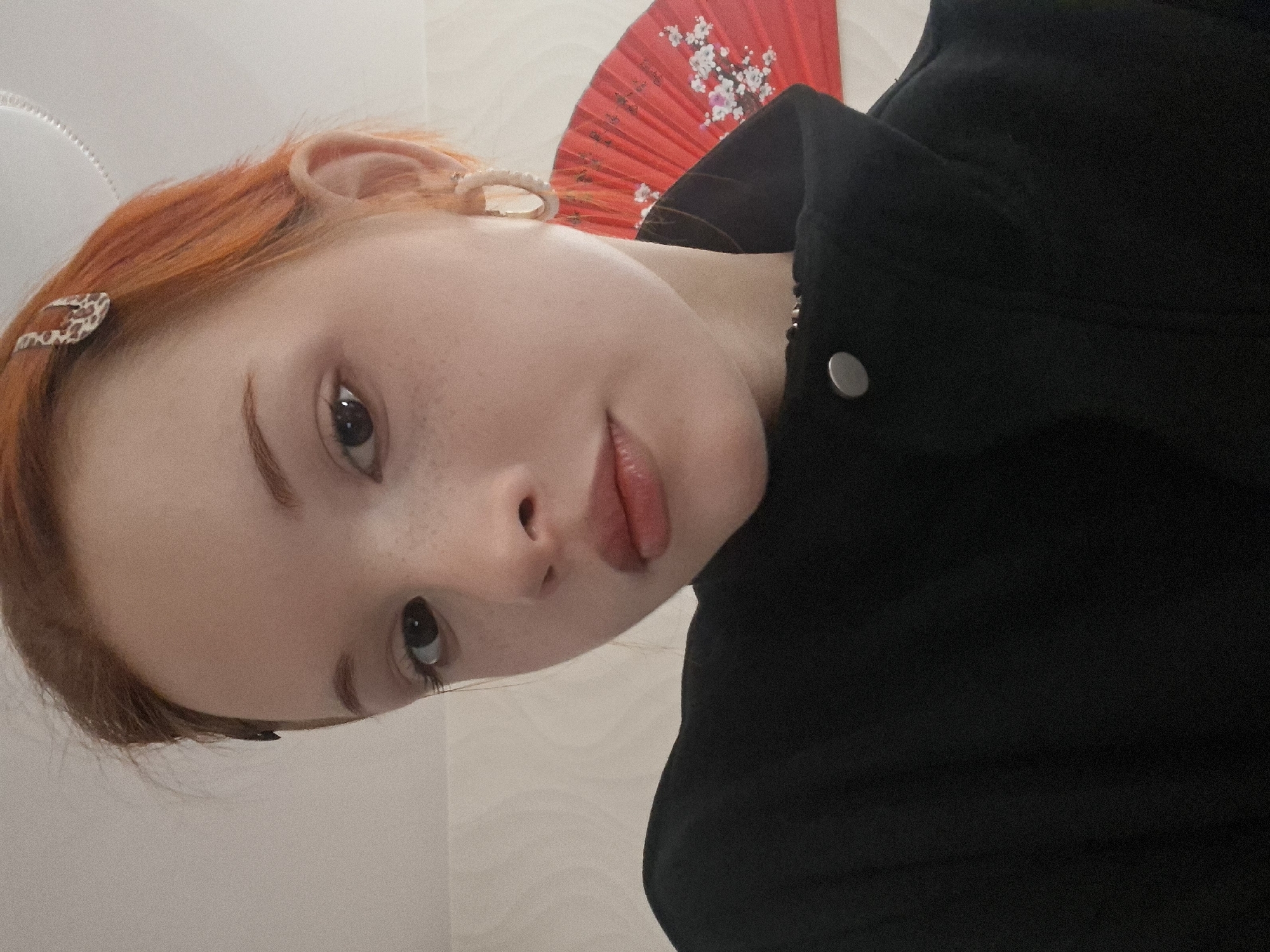 A close-up, tilted portrait captures a young individual, likely female, with vibrant orange-red hair and light skin adorned with subtle freckles. The image is notably rotated approximately 90 degrees counter-clockwise.
The subject's face is central, with dark eyes looking directly into the camera. Their eyebrows are defined, and their lips are slightly parted, displaying a natural, glossy pink hue. Several small accessories are visible: a white, possibly pearlescent, hoop earring in the right ear, and two hair clips securing strands of hair—one with a light pattern near the forehead (resembling leopard print) and another white, rounded clip further back.
Behind the person's head, a bright red folding fan, decorated with a white cherry blossom pattern, is partially visible against a light-colored wall. The individual is wearing dark, possibly black, clothing or a jacket, with a single silver button visible on the right side of the frame. The background is plain and light, suggesting an indoor setting without specific location cues. No visible text is present. The subject maintains a calm and composed expression.