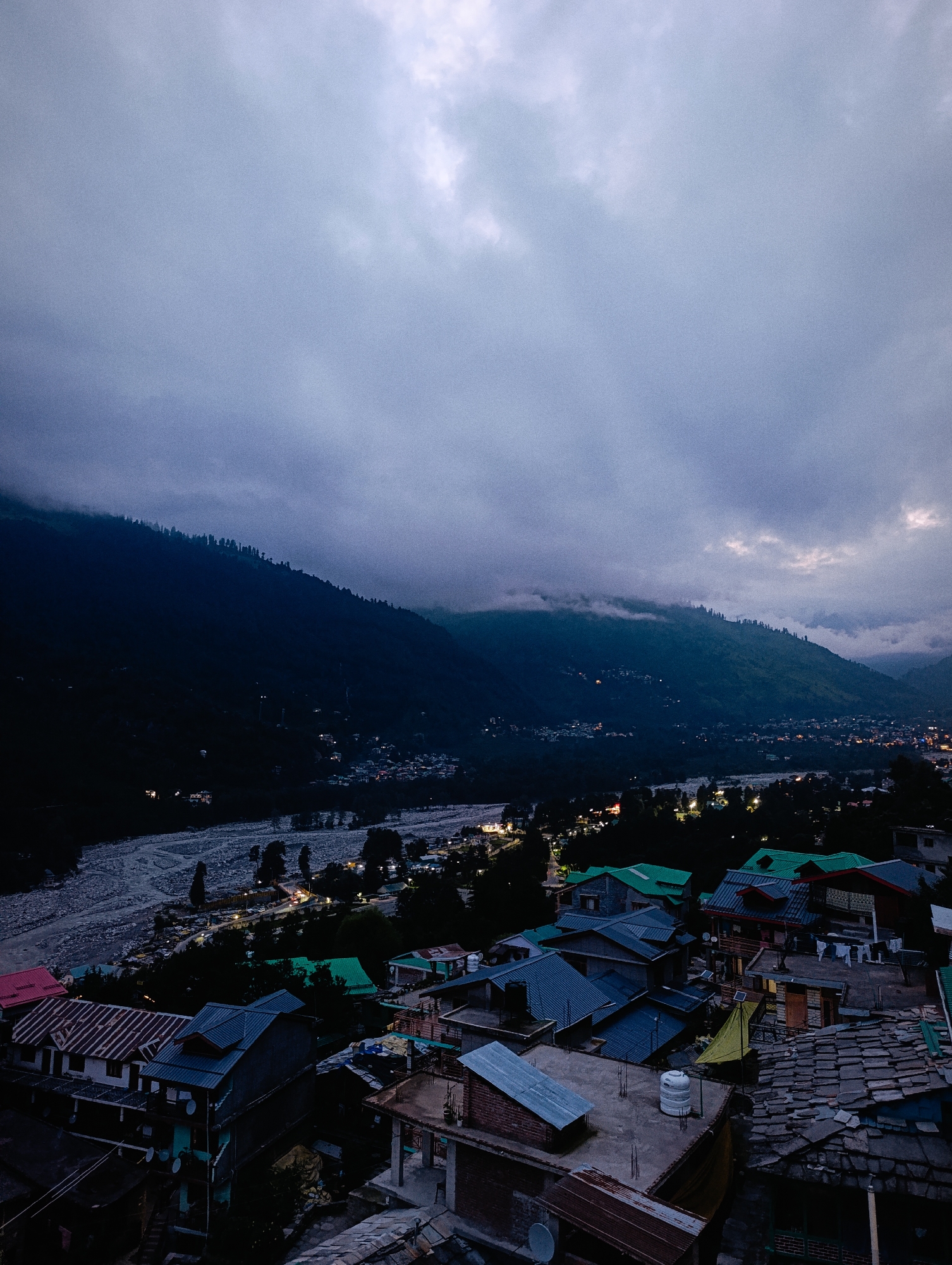 The image depicts a twilight scene of the town of Vashisht, India, nestled within a mountainous valley. The foreground is dominated by a cluster of houses with varied roof structures, including slate, corrugated metal, and brightly colored green and pink asphalt shingles. Some buildings feature satellite dishes and water tanks on their rooftops. A riverbed, appearing rocky and wide, runs through the middle ground, with a road running alongside it, illuminated by streetlights. Further back, the town lights twinkle as they spread across the valley floor, against the dark, tree-covered slopes of the surrounding mountains. The sky is overcast with heavy, dark clouds, tinged with hints of diffused light. The overall atmosphere is one of approaching night in a rural mountain town.