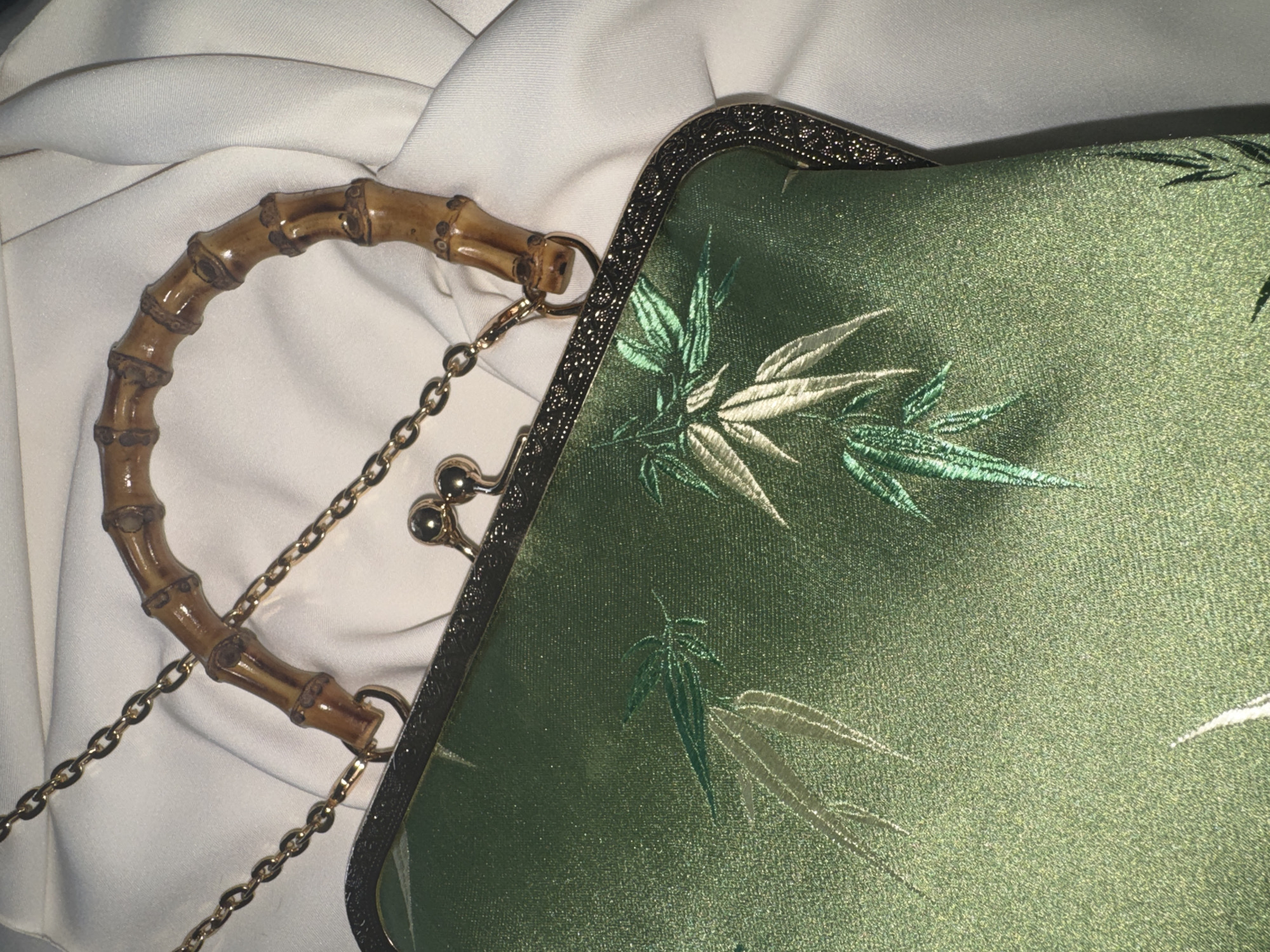 A close-up shot captures a green silk clutch purse with intricate bamboo leaf embroidery. The purse features a bamboo handle and a gold-colored chain strap. The clasp is adorned with two small gold spheres. The purse rests on a draped white fabric, suggesting a soft, luxurious setting. The lighting is dim and focused on the purse, creating a dramatic effect. The background is blurred, emphasizing the details of the purse. The overall impression is one of elegance and vintage charm.