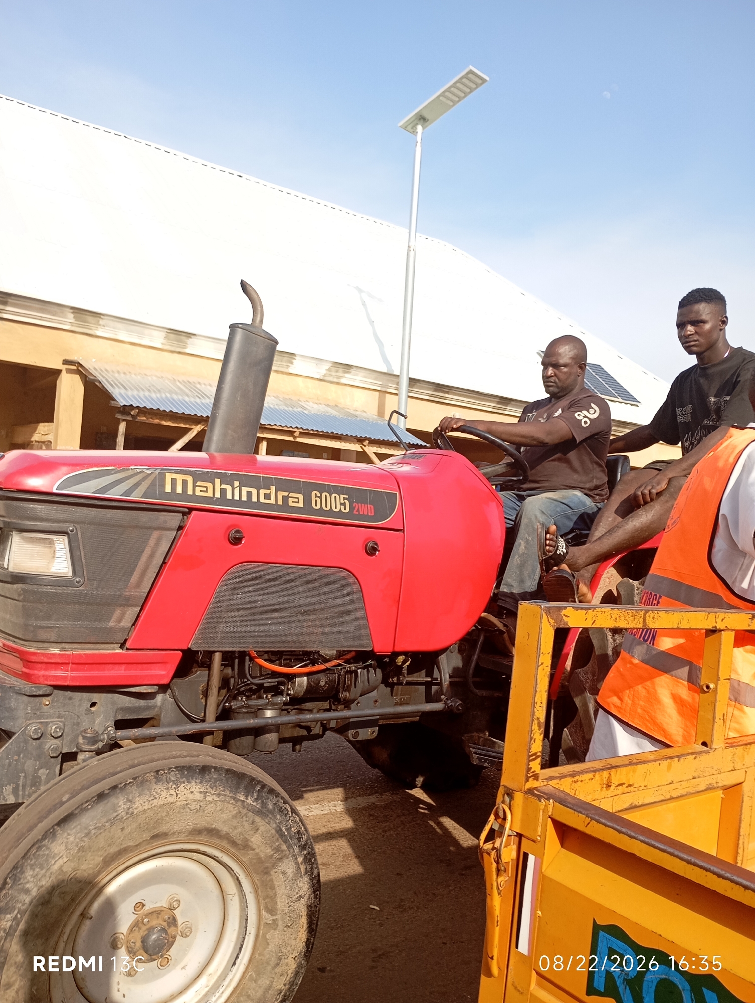 In Shendam, Nigeria, two men are on a red Mahindra 6005 2WD tractor. The man in the driver's seat is wearing a brown t-shirt and blue jeans, and is operating the tractor. The second man is seated behind him, wearing a black t-shirt. Next to the tractor is a yellow trailer with orange safety vests visible inside. In the background, there is a building with a white roof and a solar-powered street lamp. The sky is clear and blue.