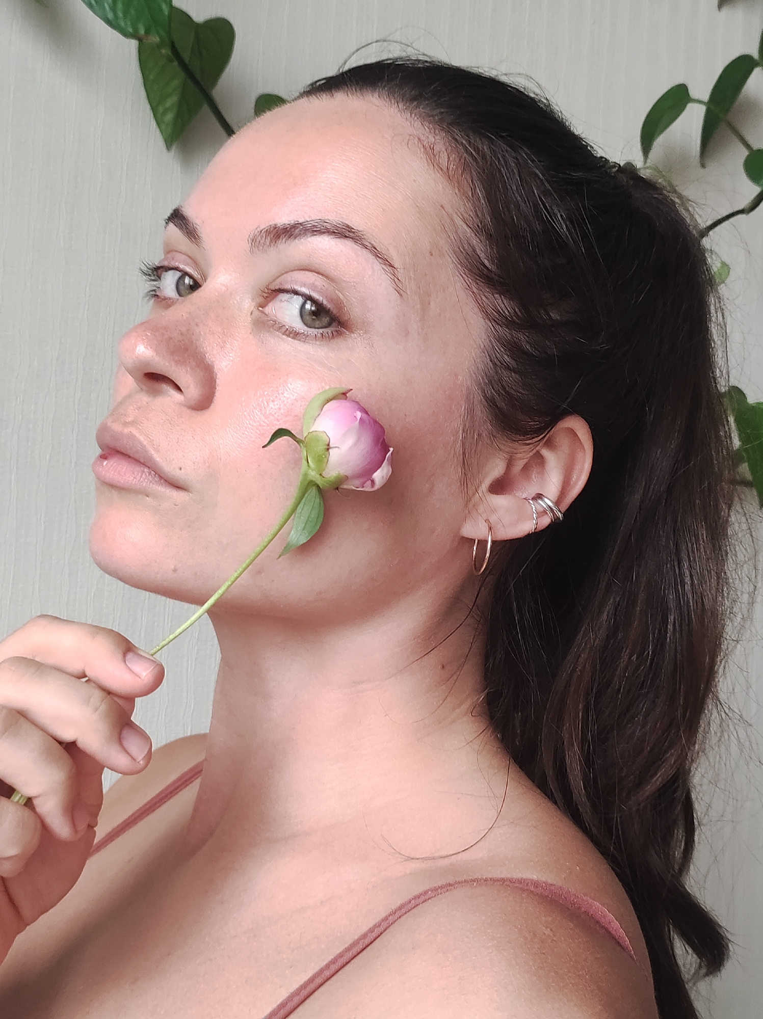 A medium shot shows a young woman with fair skin, brown hair pulled back in a ponytail, and brown eyebrows. She is holding a pink peony bud with a green stem and leaves near her right cheek. Her right ear has a small hoop earring and a silver ear cuff. The background is a light-colored textured wall, with green leaves from a plant visible in the upper left and right corners. The woman is wearing a pink camisole. The scene is an indoor setting, possibly a home. There is no identifiable location in the city of Chaikovsky, Russia.