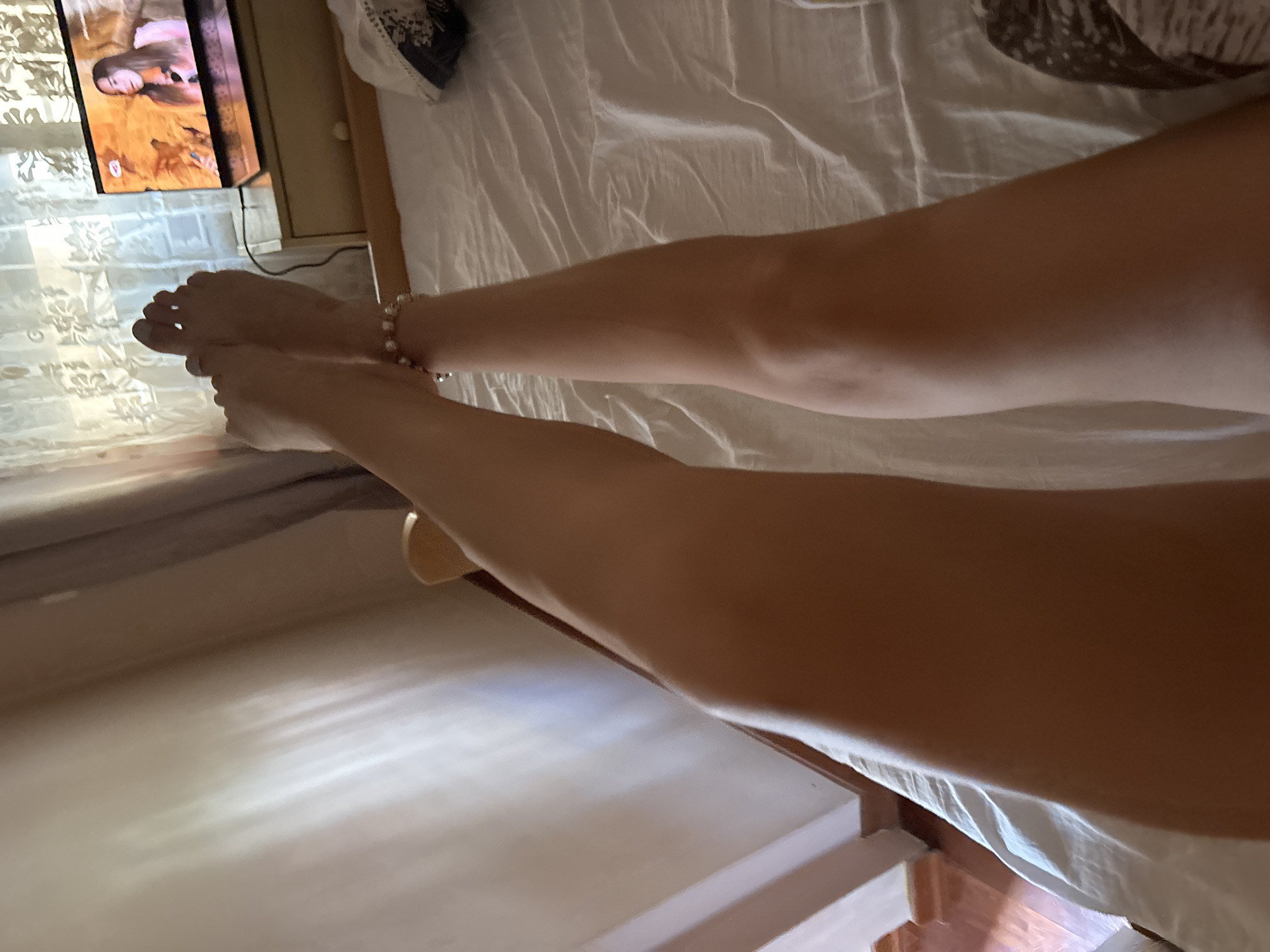 The media file features a close-up view of a person's legs and feet resting on a white, rumpled sheet, likely a bed. The person is wearing an anklet made of small, pearl-like beads. The legs are tanned and smooth, suggesting a daytime setting. To the left, a television screen is visible, displaying an image of a woman with long hair. The room appears to be dimly lit, with sunlight casting striped shadows on the floor and wall in the background. The overall impression is one of relaxation and leisure. The context provided indicates the location as Budva, Montenegro, which aligns with the warm lighting and casual setting. No text is clearly legible in the image.