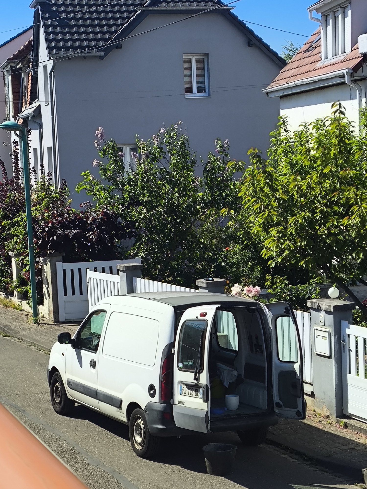 A white Renault Kangoo utility van is parked on a street in Hoenheim, France. The van's rear doors are open, revealing cleaning supplies inside, including a bucket. A dark bucket sits on the street near the rear of the van. The van is parked next to a light gray residential building with a dark roof and a white trim. A white picket fence and gate border the property. The adjacent building is light pink with a red roof. Trees and bushes are in the background. The license plate of the van is visible: FJ-112-DL.