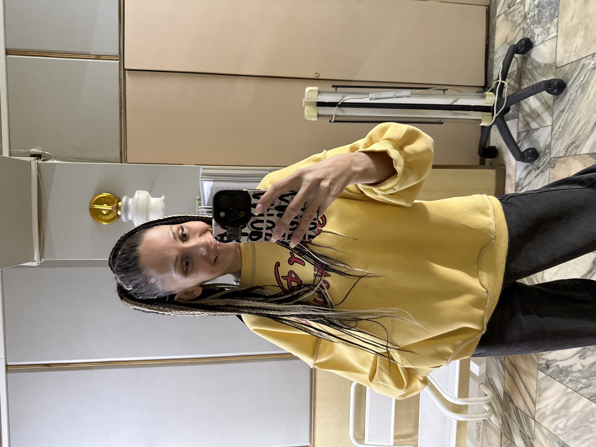 A woman is taking a mirror selfie indoors. She is standing and looking towards the camera (reflected in the mirror) with a slight smile. She has long, dark and light-blonde braided hair, some of which falls over her left shoulder and down her front. She is wearing a bright yellow, long-sleeved sweatshirt, which appears to have an indistinct black and red graphic design on the front. Her lower body is clad in dark-colored pants, likely jeans. She holds a black smartphone with a patterned case in her right hand, positioned to capture the selfie. Her fingernails have a light polish, possibly with a subtle design.
The background shows a room with various elements. To the left, there are two-toned wall cabinets or wardrobe doors, one section light grey and another light beige/brown, separated by thin gold-colored trim. Below these, a golden and white decorative fixture is visible. On the right, part of a black office chair base with wheels is seen, along with a light-colored bar or fixture behind it. The floor is covered in large, polished tiles with a mottled grey and white, marble-like pattern. A white metal chair or stool with a mesh seat is partially visible in the lower right foreground, with the word "Sorgojes" faintly legible on its frame.