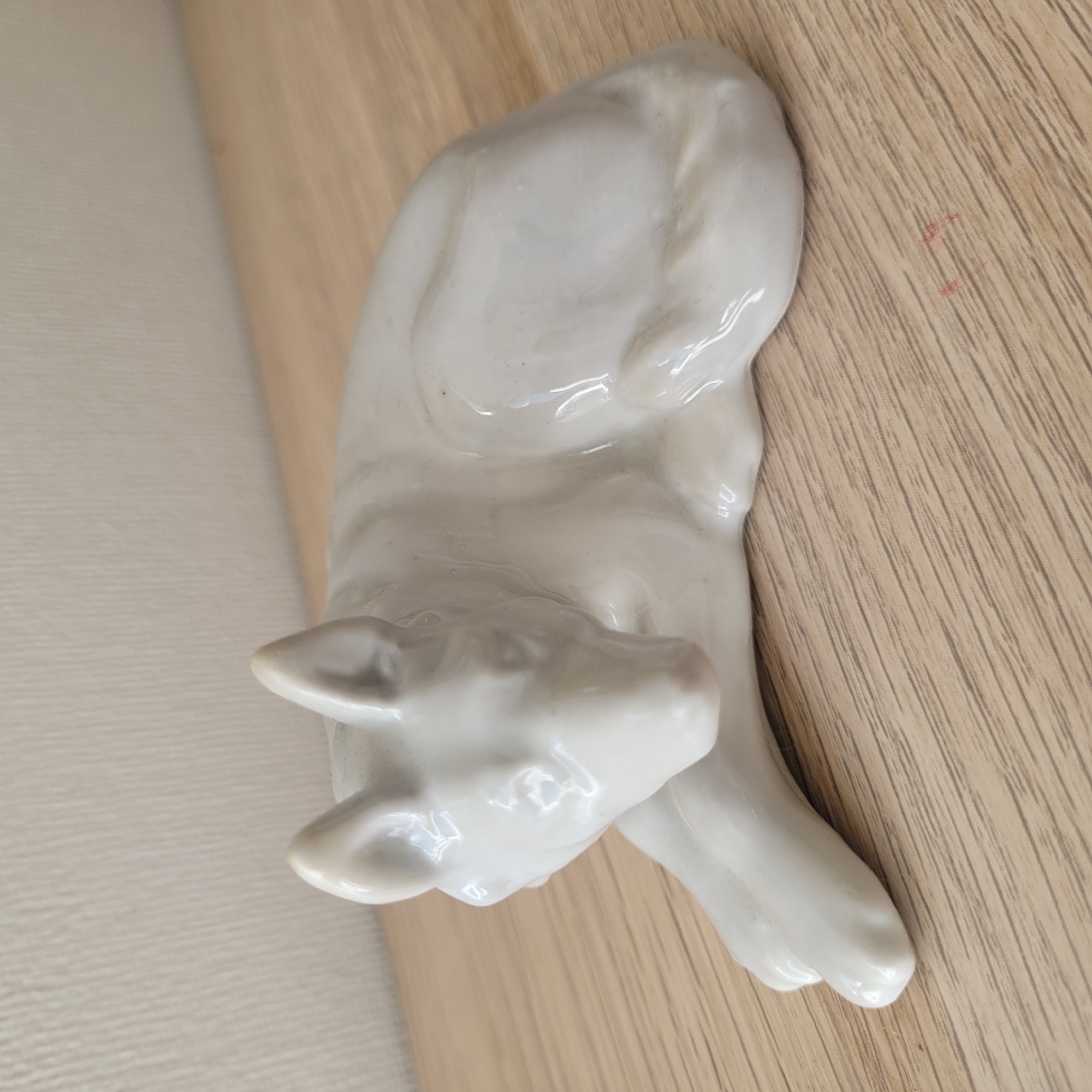 A white ceramic figurine of a dog is positioned on a light wood-grain surface, which appears to be a table or shelf. The figurine is lying down, with its head turned to the left and its tail curled around its body. The background is a textured, off-white wall. There are no people or any other objects visible in the scene. The image does not provide any information about the location in the city of Jūrmala, Latvia.