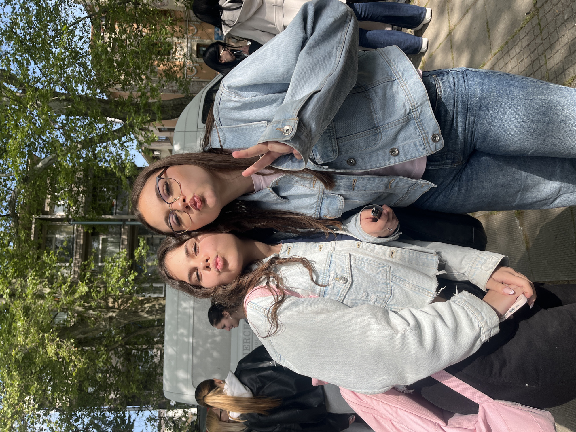 This image captures two young women posing playfully for a photo in an outdoor urban setting in Plovdiv, Bulgaria, during what appears to be a bright, sunny day. The photograph is oriented horizontally, showing the subjects rotated 90 degrees counter-clockwise.
In the foreground, one woman with long dark hair, wearing glasses with thin gold frames, a light pink top, a light blue denim jacket, and dark jeans, poses with a peace sign made with her left hand and puckers her lips in a "duck face." A ring is visible on her right hand. Beside her, another woman with long brown hair, wearing a similar light blue denim jacket (appearing slightly distressed) over a pink top or strap, and dark trousers, also makes a "duck face." Her hands are clasped in front of her, holding a light pink mobile phone, and her nails feature French tips. A pink bag strap is visible on her left side.
The background features lush green trees and a clear blue sky on the left, with glimpses of buildings and windows beyond the foliage. On the right, a white van is partially visible, bearing partial text that seems to read "MERCE". Further back, several other individuals are visible, some wearing lighter coats and a black leather jacket. The ground is paved with light-colored blocks, and the presence of shadows indicates bright sunshine. The overall scene conveys a casual and friendly atmosphere.