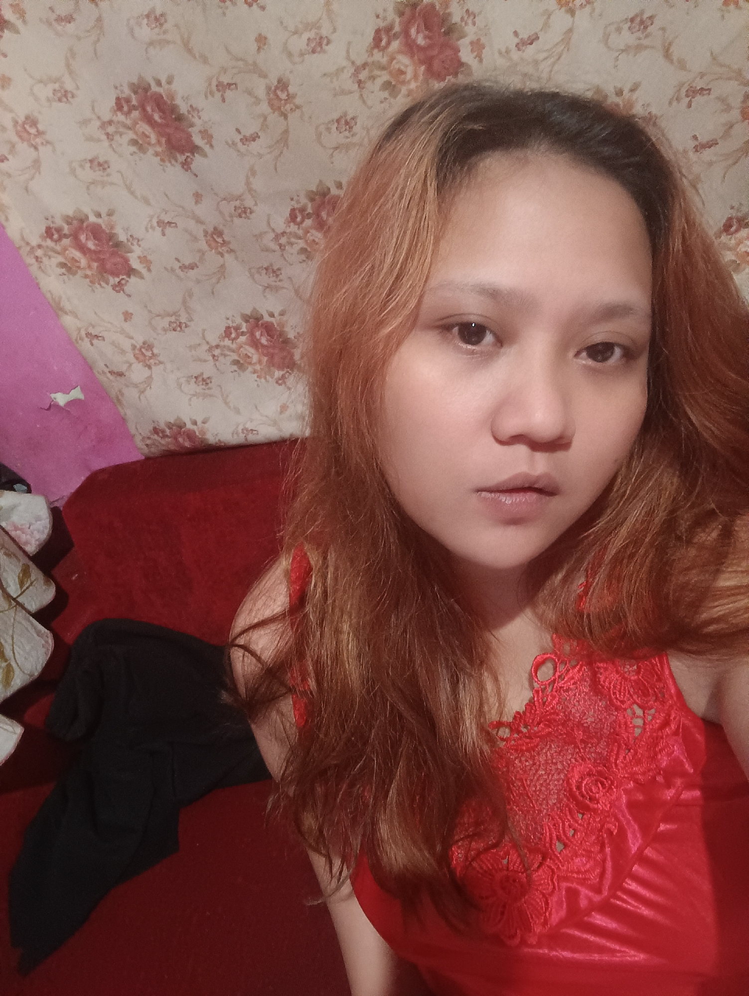 A young woman with reddish-brown hair and light skin poses for a selfie indoors. She is wearing a red satin nightgown with lace detailing on the neckline. The background consists of a floral patterned wallpaper and a red couch. The lighting suggests an indoor environment, possibly a bedroom or living area. The woman's facial expression is neutral, and her gaze is directed at the camera. She is positioned slightly off-center, with her hair falling around her face and shoulders. The image appears to be taken from a close-up perspective, focusing on the woman's upper body and face. No specific location in Depok, Indonesia can be identified from the visual information provided.