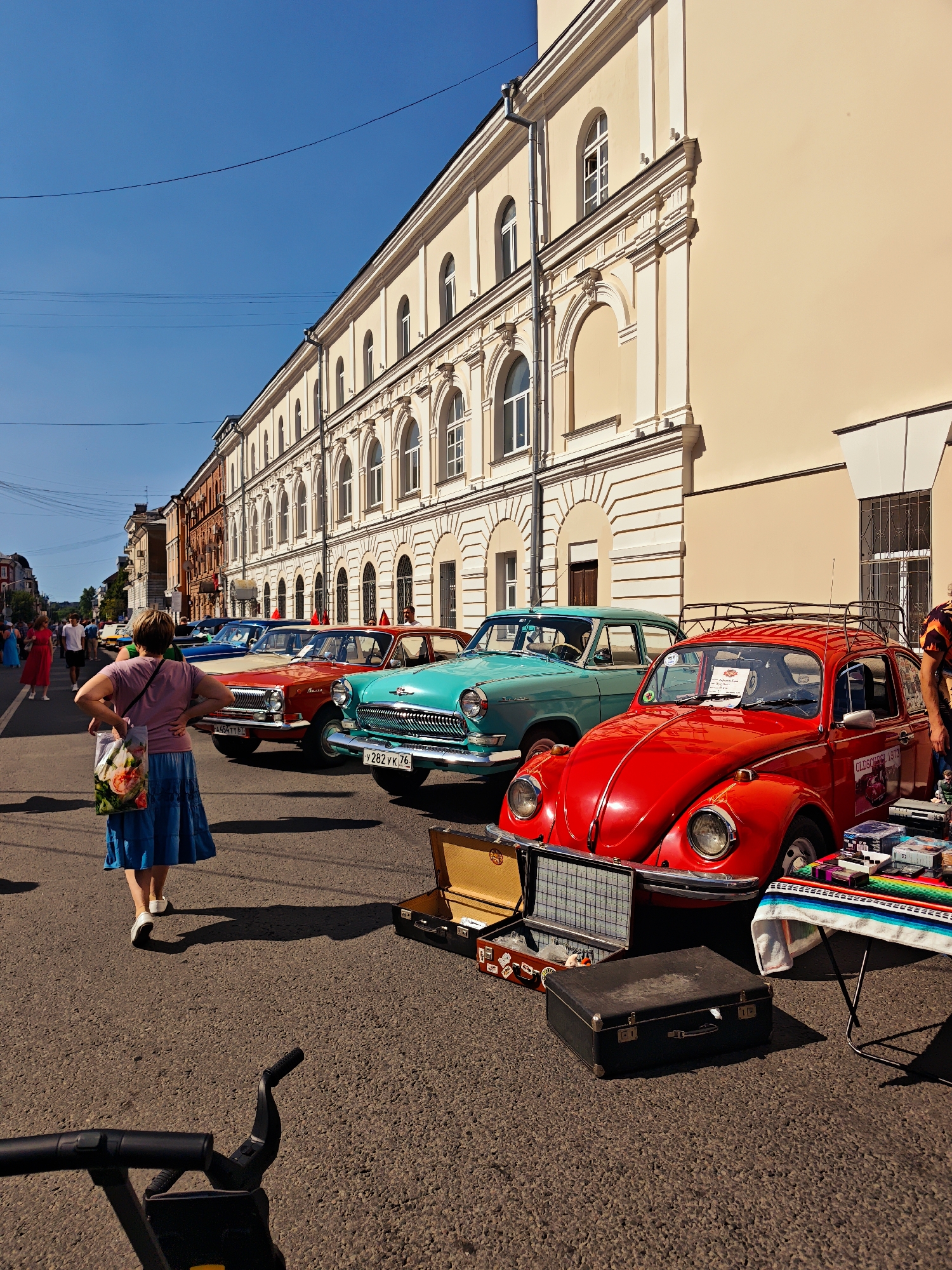 An outdoor scene on a sunny day in Tver, Russia, depicts a street lined with vintage cars and a large, light-colored building with arched windows. In the foreground, a woman wearing a pink shirt and blue skirt walks away from the camera, carrying a patterned tote bag. Several classic cars, including a red Volkswagen Beetle and a turquoise sedan, are parked along the street. Some of the cars are displaying items on tables or in open trunks, suggesting a possible car show or market. In the background, other pedestrians and buildings are visible.