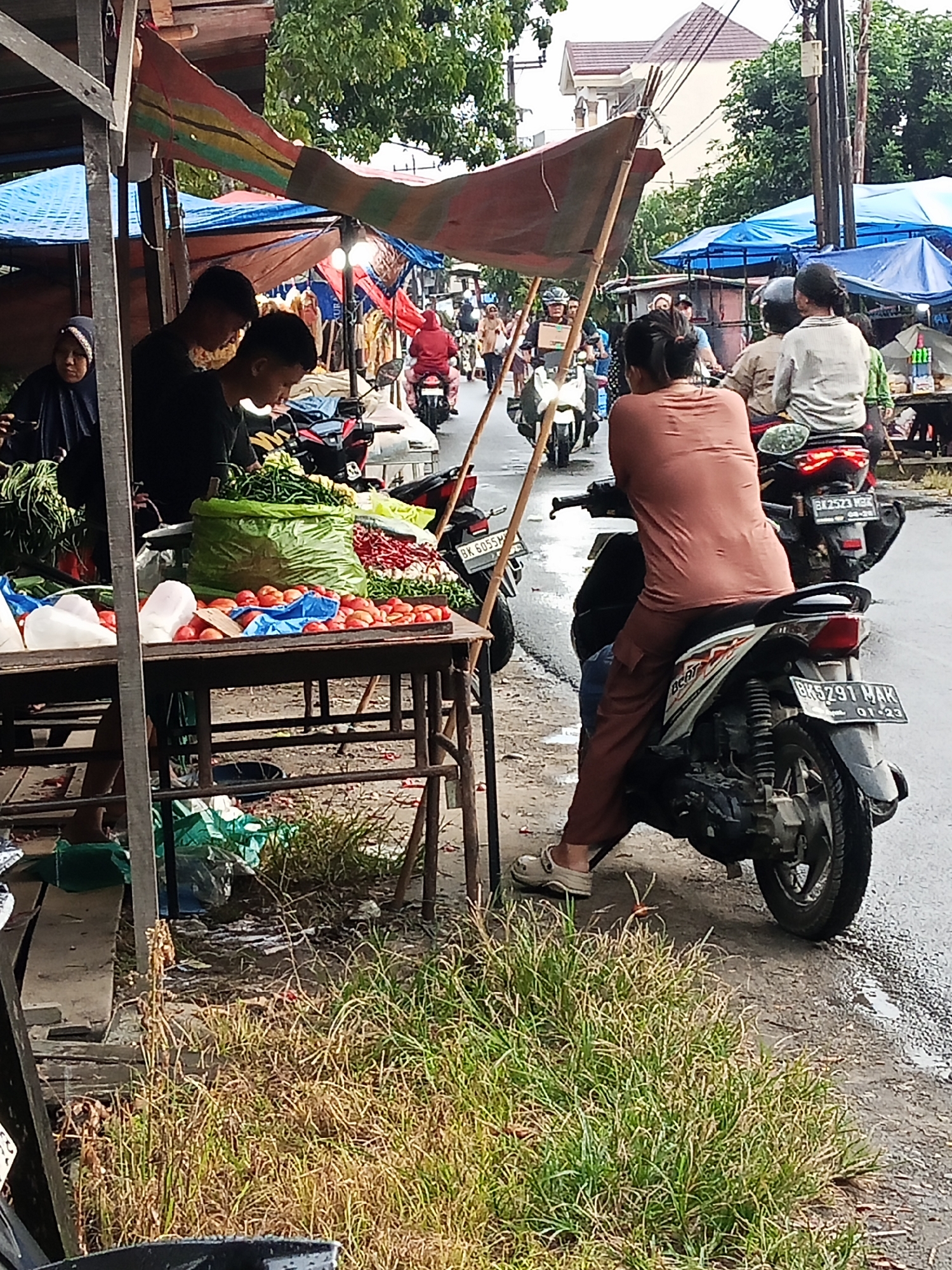 A street market scene in Wono Sari, Indonesia, captures a moment amidst the movement and activity of vendors and customers. In the foreground, a stall displays an abundance of fresh produce, including piles of red tomatoes and a large green bag filled with chili peppers and long green beans. A woman in a dark hijab stands behind the stall, her attention focused on the items. Two young men are also present at the stall, one in a black t-shirt and the other in a black shirt, appearing to assist with the produce.
Beyond the stall, the street is lined with more market stalls, some covered with blue and red tarpaulins. Several motorcycles are parked and in motion, indicating the common mode of transportation in the area. One motorcycle in the mid-ground carries a woman wearing a brown t-shirt and pants, with another person riding behind her. Another motorcycle in the foreground shows its rear wheel and license plate, which reads "BK 5291 MAK 01-23". The street appears to be wet, suggesting recent rain or damp conditions. In the background, more market activity and structures are visible, along with trees and buildings. The overall atmosphere is that of a bustling local marketplace.