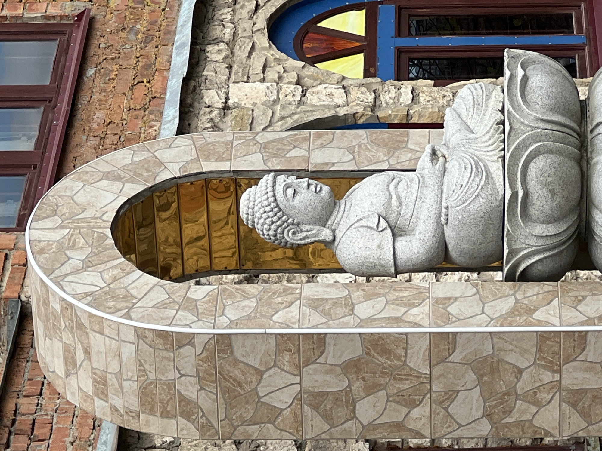 The image depicts a reclining Buddha statue situated on a tiled balcony. The statue is made of gray stone, and the background is a building with brick and stone construction, including windows with red and blue frames. The location is Kazan, Russia.