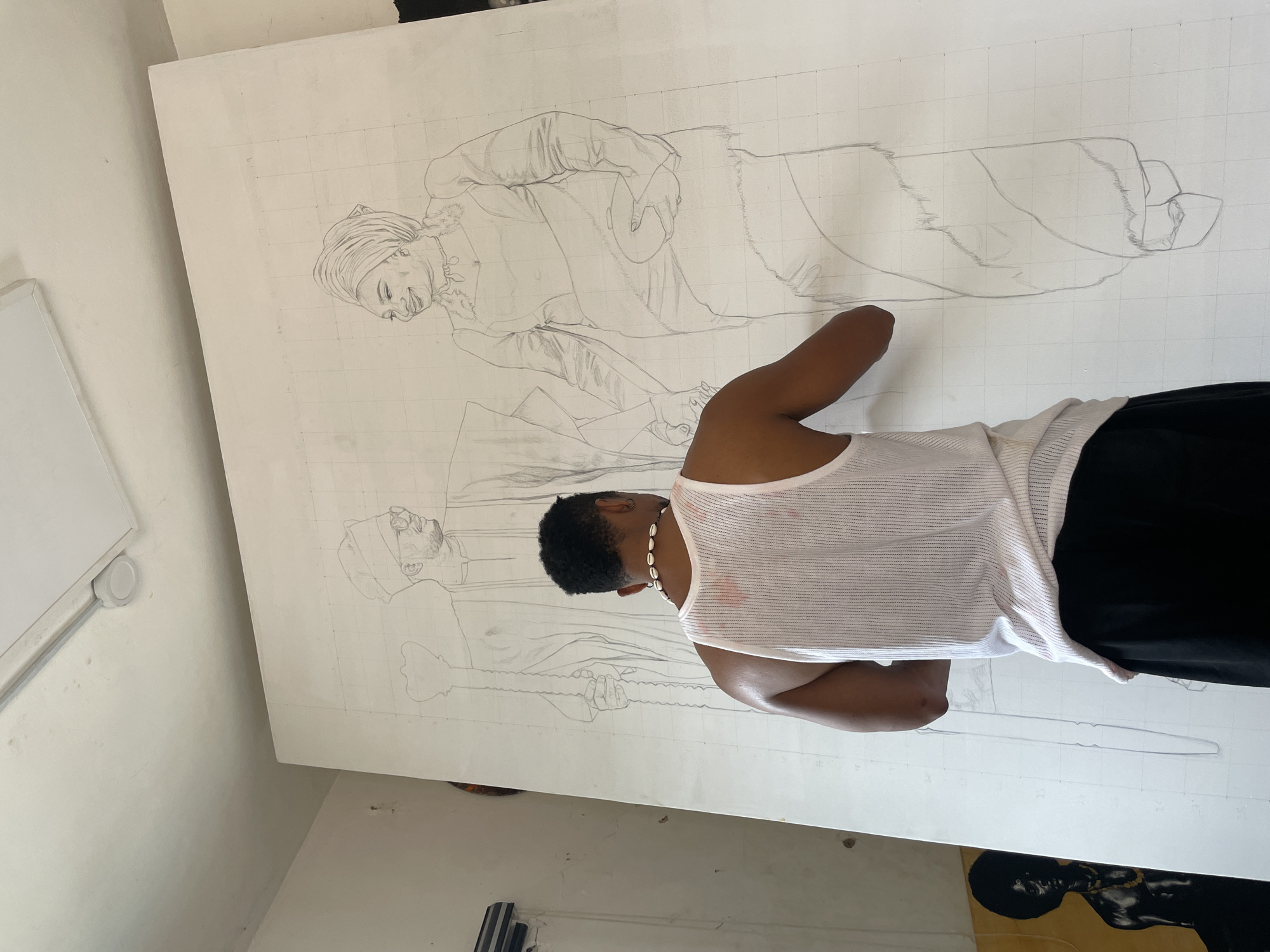 A person is sketching a large drawing on a gridded canvas in an indoor setting. The person is facing the canvas and appears to be actively drawing. They are wearing a white tank top and black pants, with a necklace of shells around their neck. The drawing depicts two figures, seemingly portraits of individuals in traditional clothing. The canvas is large, almost the size of a wall, and the grid lines are faintly visible. The room appears to be a studio or workspace, with white walls and some art supplies and other canvases visible in the background. The location is Unknown, Unknown.