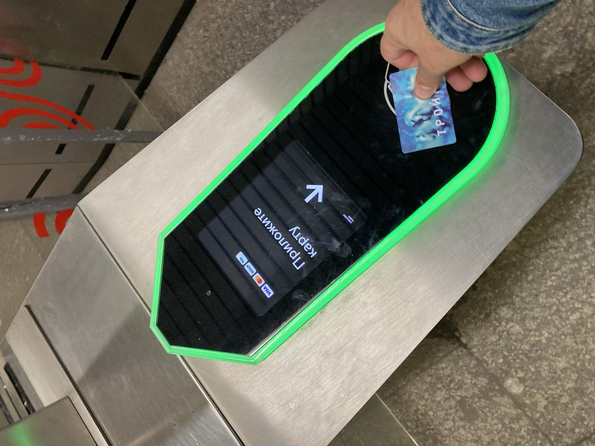 An individual, wearing a blue denim jacket, is holding a blue and white transit card labeled "ТРОЙКА" up to a turnstile reader. The reader is an elongated, hexagonal device with a black screen surrounded by a bright green light. The screen displays the Russian text "Приложите карту" (Apply card) and an arrow pointing downwards, indicating where to tap the card. Above this text, logos for Visa, Mastercard, and MIR are visible, suggesting payment options. The turnstile itself is made of brushed metal, and the surrounding environment appears to be a subway station in Moscow, Russia, as evidenced by the "ТРОЙКА" card, which is a Moscow's unified social card for public transport. The floor is a speckled grey material, and the background shows part of a metro station wall with red graphic elements and metallic poles.