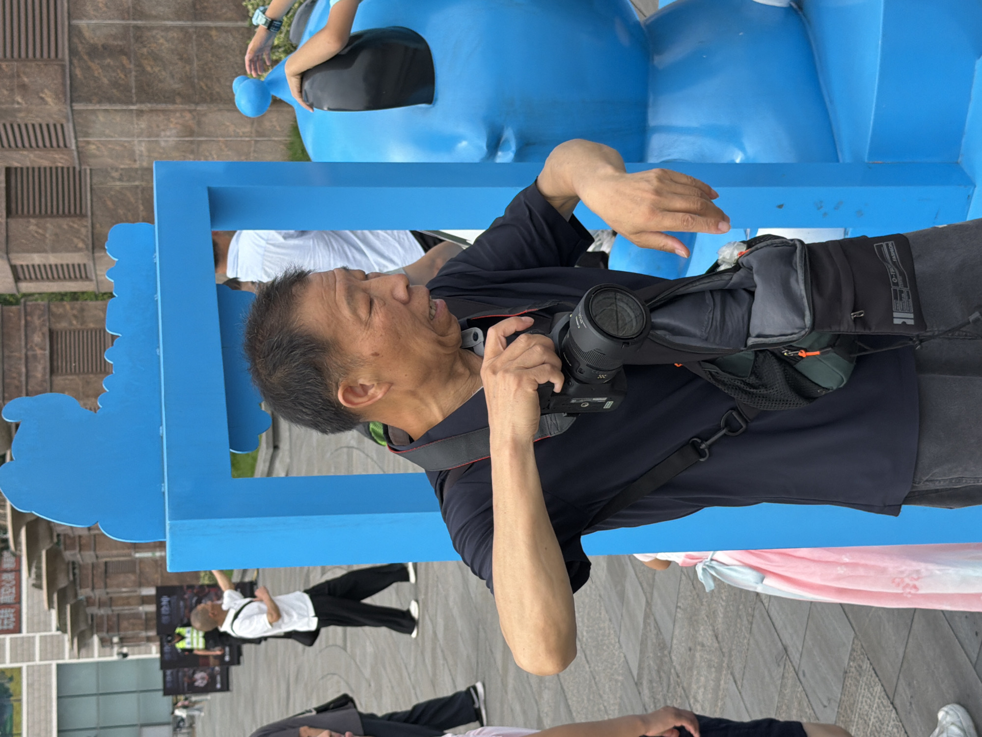 An East Asian man in a black polo shirt and gray shorts holds a black DSLR camera in his right hand, aiming it towards the viewer. He has a black strap around his neck, attached to a black and gray bag slung across his chest. He stands in front of a bright blue, abstract sculptural installation with a large rounded head, possibly a character or mascot. In the background, out of focus, are buildings with arched windows and a paved walkway. To the left, another man in a white polo shirt and black pants walks away from the camera, while a person in a yellow vest stands further in the background. A child in a pink dress is partially visible to the left of the main subject. The scene appears to be an outdoor urban public space in Yuzhong District, China.