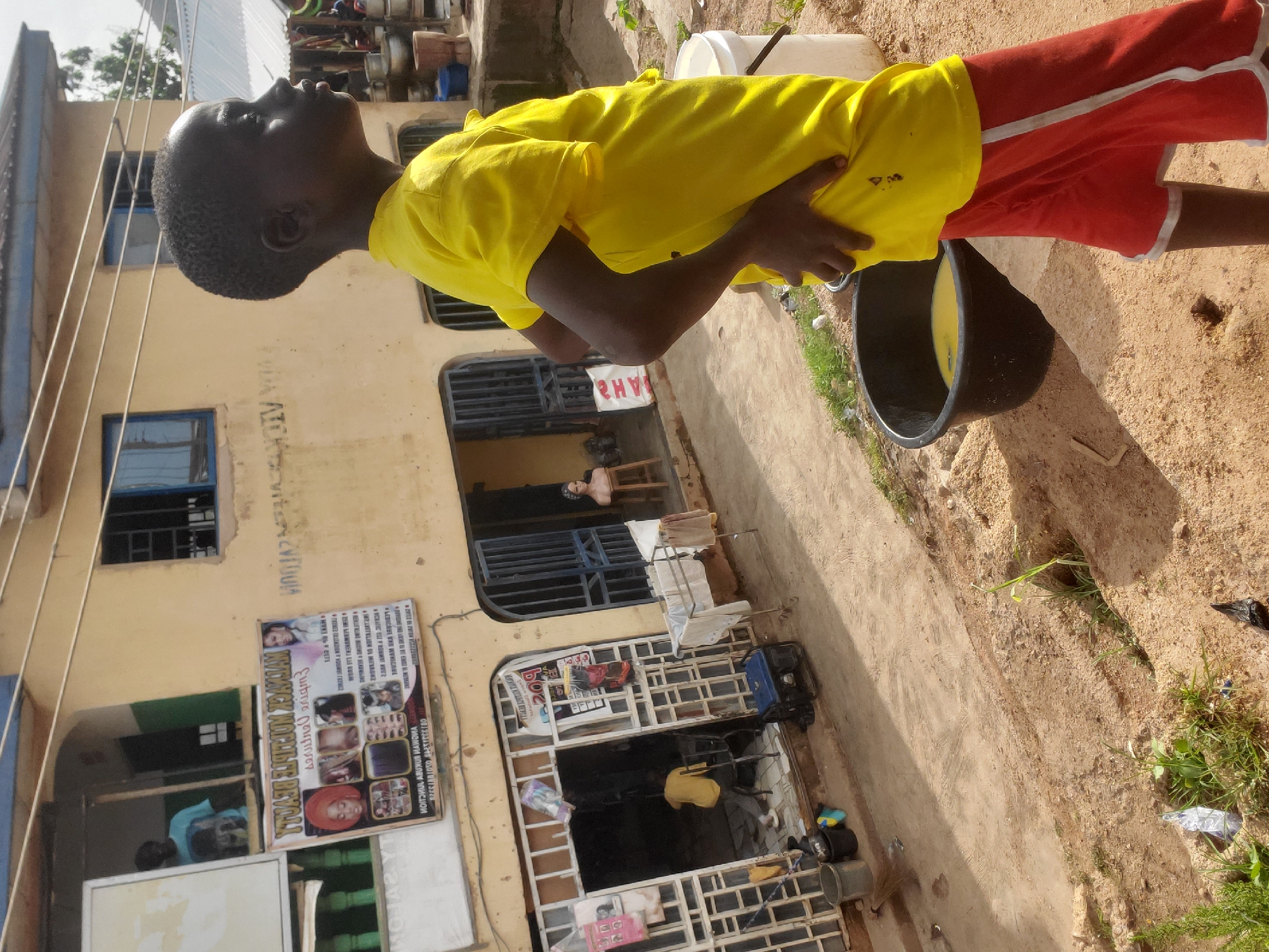A young boy wearing a yellow t-shirt and red shorts stands in a dusty, outdoor area. He is looking upwards with his head tilted back, his right hand resting on his stomach and his left hand on his hip. Beside him, to his right, is a large black bucket containing some yellow liquid, possibly water.
The setting appears to be a street or an alleyway in front of a two-story building with a pale yellow facade. The building has several windows and doors, some of which are barred. There is a sign visible on the building with text and images, featuring hairstyles and portraits. Laundry hangs on a rack near one of the upper windows. The ground is predominantly sandy and dirt.
The overall scene suggests a warm, possibly sunny day, given the brightness and shadows. The environment looks somewhat weathered and perhaps sparsely populated. There's a general sense of everyday life in a developing area.
Visible text includes:
- On the sign: "HAIR STYLE MONIQUE BELLA" and other text in a language that appears to be a local Nigerian language, likely Igbo or Yoruba, given the location context of Gwong, Nigeria.
- On a white bag: "AH2" (reversed).