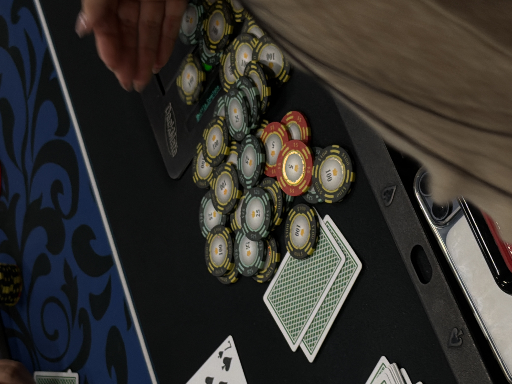 An overhead, slightly angled shot captures a portion of a card game, likely poker, taking place on a black felt table. A person's hand, with manicured nails, reaches into the frame from the upper left. A significant pile of poker chips is scattered across the table. These chips are primarily yellow/gold ($100 denomination), light blue/green ($25 denomination), and red ($5 denomination). Many chips feature a crown symbol in their center.
To the right of the chip pile, a stack of green-backed playing cards lies face down, with another card placed on top. In the lower left, two face-up cards are visible, one clearly identifying as the 7 of Spades. A "DeckMate" card shuffler/dispenser device is positioned behind the main cluster of chips. On the right side of the table, a smartphone in a dark case, resembling an iPhone, is partially visible, with a small spade symbol embossed on its case.
The scene suggests an active game due to the scattered chips and cards. Visible text includes "100", "$100", "25", "$25", "5", "$5" on the chips, "DeckMate" on the shuffler, and "7" on one of the playing cards. The indoor setting and artificial lighting indicate either a casino environment or a home game setup.