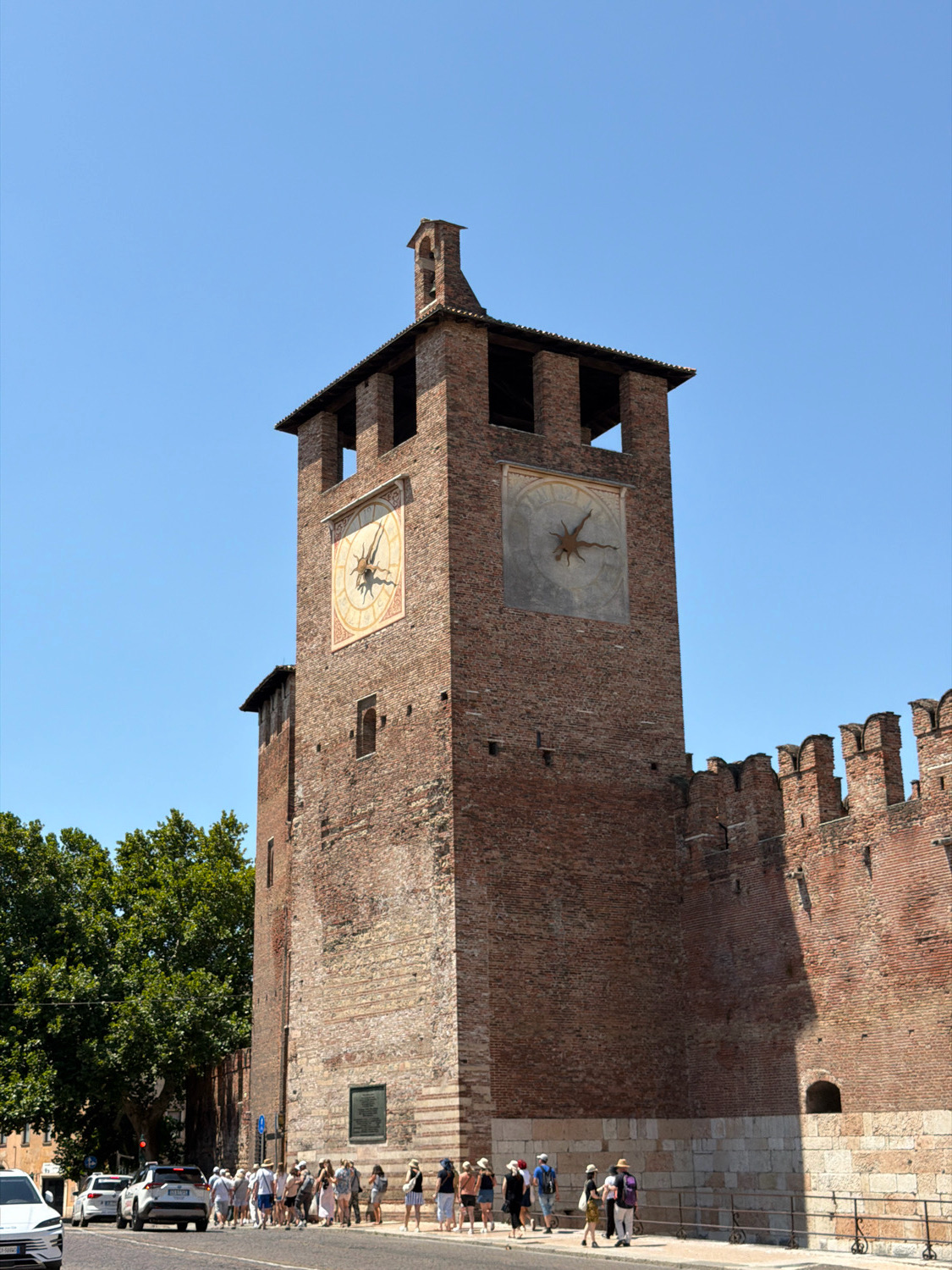 The image shows the exterior of the Torre dei Lamberti in Verona, Italy, a tall medieval brick tower with two clocks on its facade. The tower is part of the city's historic center and stands adjacent to a long brick wall with battlements. A clear blue sky forms the background. In the foreground, a group of tourists is gathered on a sidewalk next to a street with several cars. Trees are visible to the left of the tower, and a small plaque is mounted on the tower's lower section. The scene depicts a sunny day with distinct shadows cast by the tower and wall.