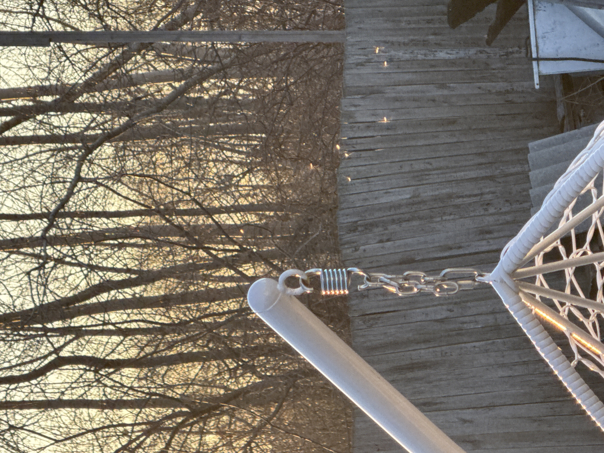 The image presents a partial view of a white hammock chair suspended by a metal chain and spring from an overhead support structure. The hammock's frame is white, with a rope or cord woven to create the seat. A wooden deck with parallel planks is visible behind the chair. The planks have gaps in between them. The background on the left shows leafless trees against a bright sky, possibly at sunset or sunrise. The location may be in Chaikovsky, Russia, judging by the question, but the image does not explicitly show this.