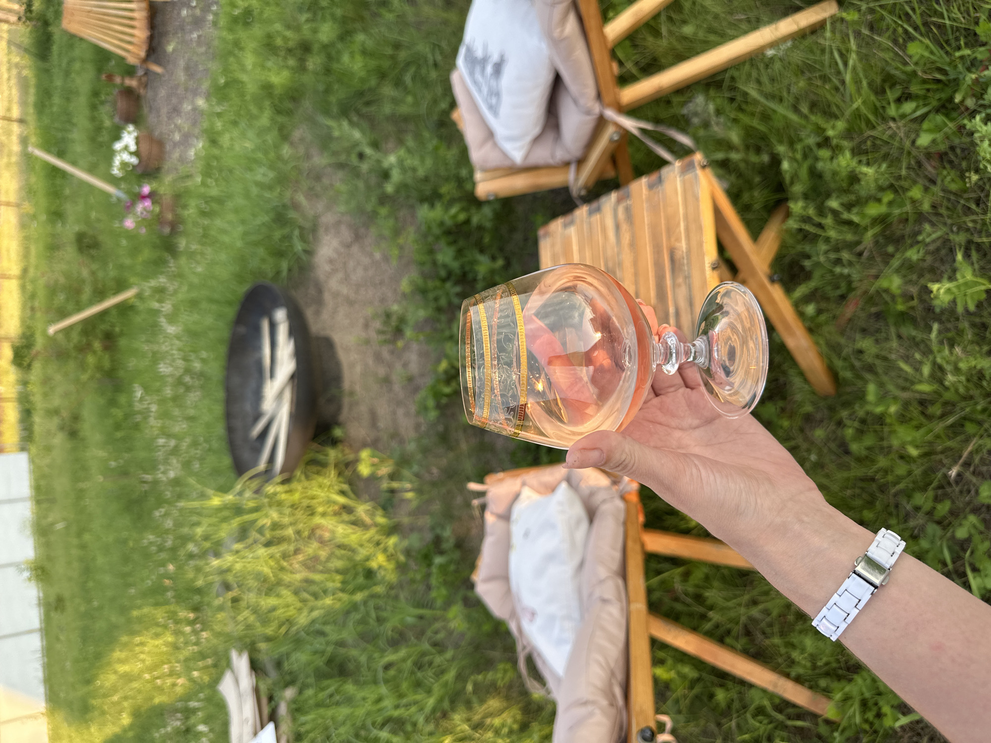 A person's hand, wearing a white watch, holds a brandy glass filled with a pink liquid. The glass has gold decorative banding on its rim. The setting is outdoors on a grassy area, with two wooden chairs with cushions and a small wooden table visible in the background. A fire pit is also present. The overall scene suggests a relaxed outdoor gathering. The location is not specified in the city of Unknown, Unknown.