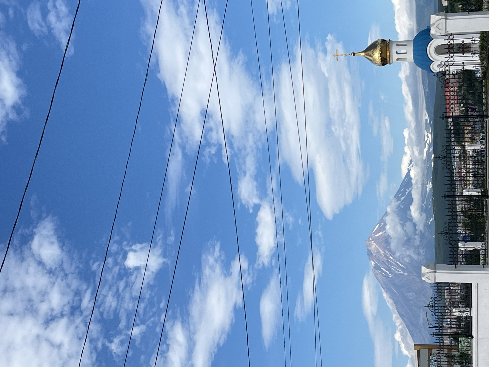 The image captures a view of Petropavlovsk-Kamchatsky, Russia, with a prominent volcano in the background and a small Orthodox church in the foreground. The volcano, likely Koryaksky or Avachinsky, is partially shrouded in clouds, with patches of snow visible on its slopes. To the right, a white church with a blue dome and a golden cross stands out against the bright blue sky dotted with white clouds. A black wrought-iron fence separates the viewer from the church and the distant cityscape, which includes red-tiled roofs. Several dark power lines crisscross the upper portion of the frame, obscuring parts of the sky. The scene appears to be set on a clear, sunny day.