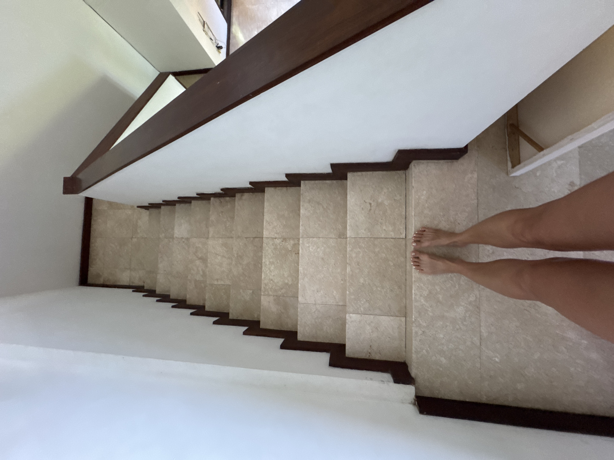 A high-angle view of a staircase with a brown wooden railing along the left side. The stairs are covered in a beige stone tile. The walls and ceilings are white. A person's legs and feet are visible on the right, standing at the top of the stairs. The location is possibly in Kuta Selatan, Indonesia, based on the architectural style often found in the area.