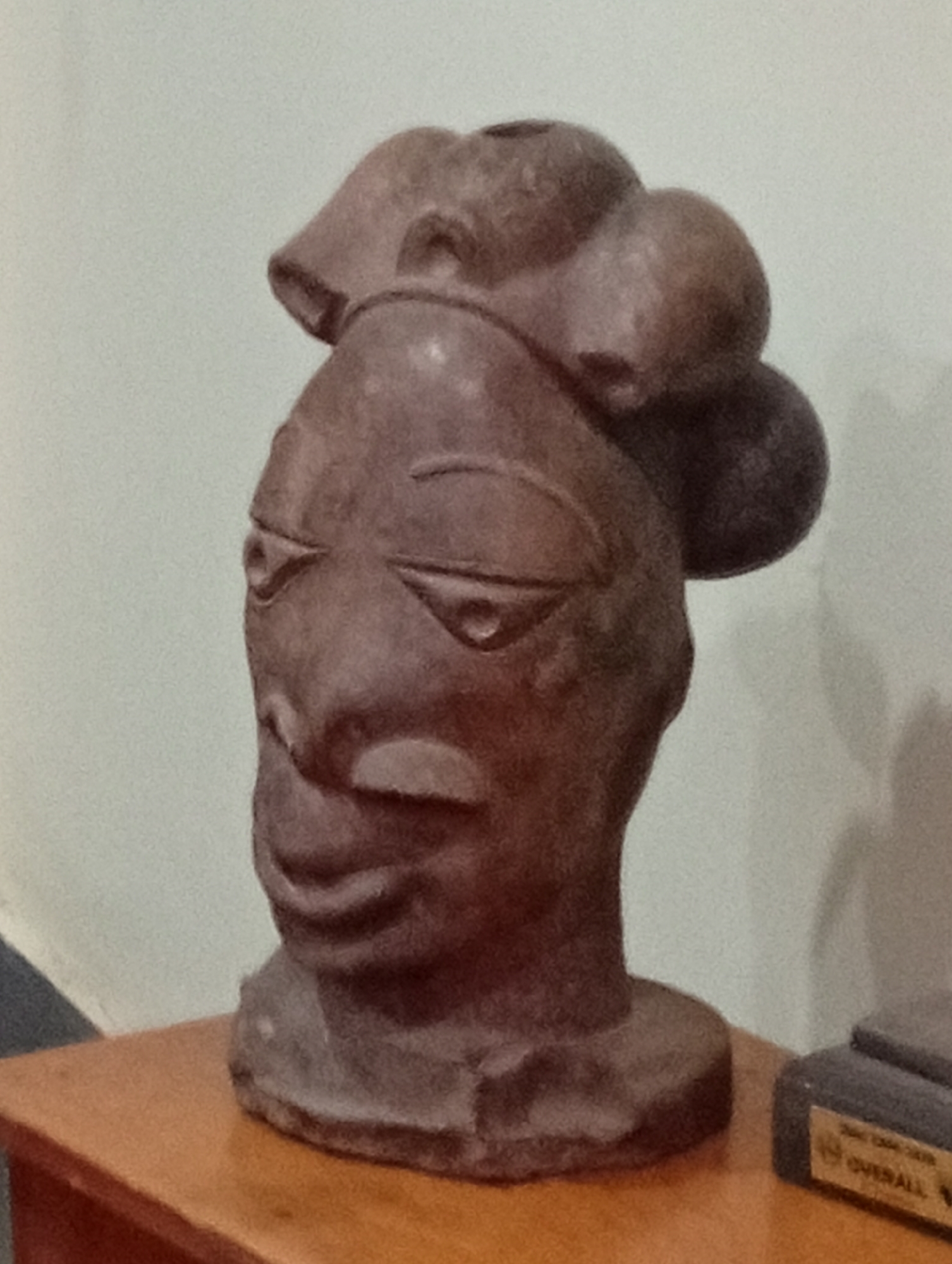 A medium shot of a brown statue of a head on top of a wooden surface, possibly a table. The statue appears to be of African descent and has several protruding rounded shapes on top of the head. The statue has eyes, nose, and mouth features that are stylized. Behind the statue is a light-colored wall. To the right of the statue is another object which could be a trophy. The scene is inside, probably in a residential location in Dadi Riba, Nigeria. There are no visible people, actions, or interactions present.
