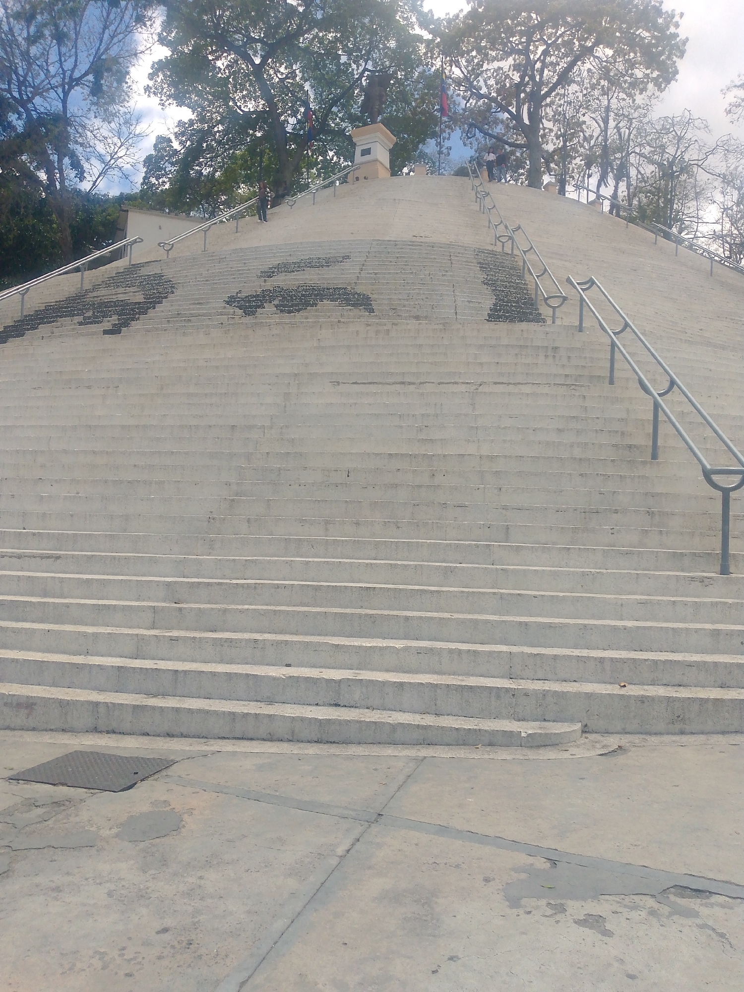 This image captures a wide, expansive concrete staircase leading up a steep incline towards a hill crowned with trees. The stairs are equipped with parallel grey metal handrails that run along both sides, extending far up the slope. Several sections of the stair risers and treads feature dark, decorative patterns, possibly mosaics or painted designs.
In the immediate foreground, a flat concrete ground surface shows signs of wear, including cracks and stains. A square metal grate or manhole cover is visible on the left side of this ground area.
As the stairs ascend, a few people can be seen at various points. Some individuals are walking on the steps, while others are standing near the top of the hill.
At the summit, lush green trees with dense foliage are prominent, contrasting with some bare branches against a partially cloudy sky. A light-colored, multi-tiered monument or structure stands near the top of the stairs, and an indistinct flag, appearing to be the Venezuelan flag with its horizontal stripes, flies from a pole nearby.
The scene is brightly lit by natural daylight, indicating it is daytime. The combination of the architecture, vegetation, and the Venezuelan flag context suggests the location is a public space or landmark in Caracas, Venezuela. No discernible text is visible in the image.
