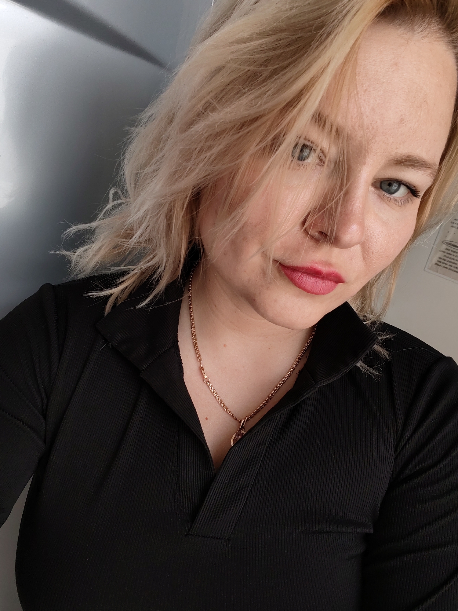 A fair-skinned woman with blonde hair is seen in a close-up shot. Her hair partially covers her face, but her blue eyes are visible. She is wearing a black ribbed shirt and a gold necklace. The background consists of a light gray surface and a white wall with a partially visible document. The image does not provide information to establish a location in Izhevsk, Russia.