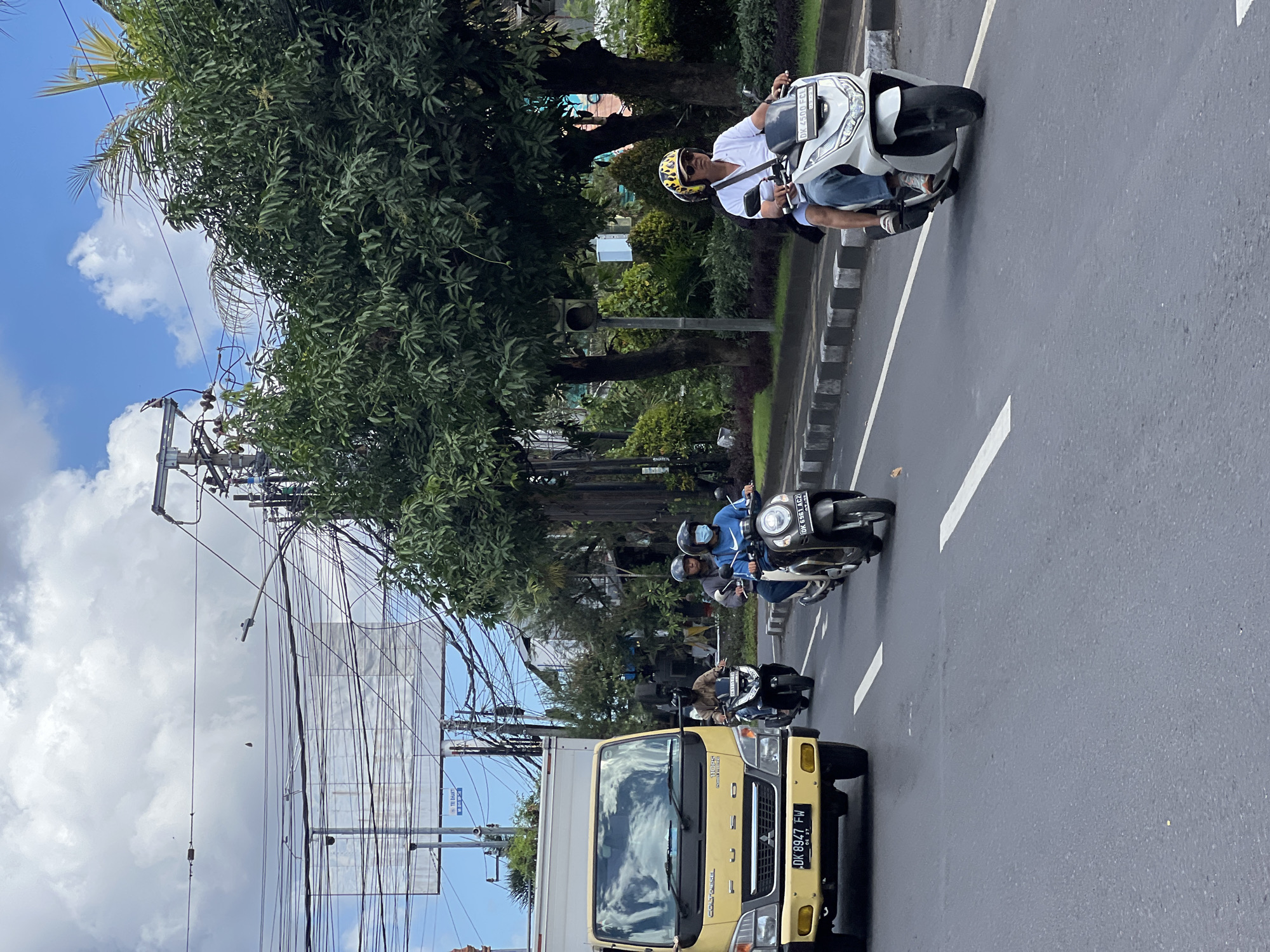 A bright, overcast day in Denpasar, Indonesia, features a bustling street scene with several motorbikes and a yellow truck navigating the road. The foreground shows the road with white lane markings. In the middle ground, a yellow FUSO truck with license plate "DK 8947 FW" is on the left side of the road. Several motorbikes are in motion. A man wearing a helmet and a blue shirt with a white Nike logo is on a white motorbike on the right, with license plate "DK 6961 AC2". Another motorbike to his left carries two people, one wearing a helmet and a blue face mask. Further left, another motorbike with a single rider is visible. The background is filled with lush green trees and electrical wires overhead, indicating an urban environment. A billboard is visible behind the trees. The weather appears to be humid.