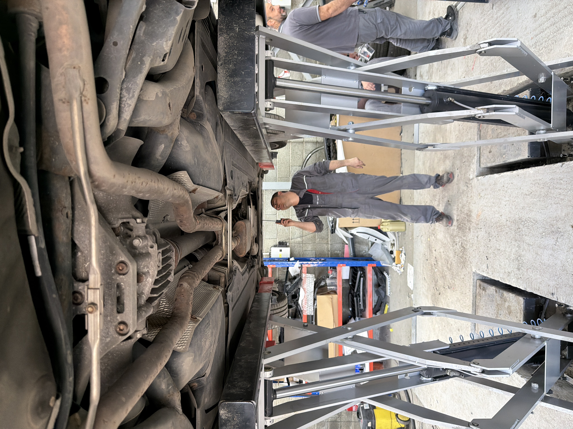 An image shows the underside of a vehicle on a lift in a garage setting, featuring two men in work attire. One man lies extended beneath the vehicle, holding a tool. The other stands adjacent to the lift, partially visible. The vehicle's exhaust system and undercarriage components are prominent. Various automotive tools and parts are scattered in the background. There is no discernible evidence to confirm the location as Burgas, Bulgaria.