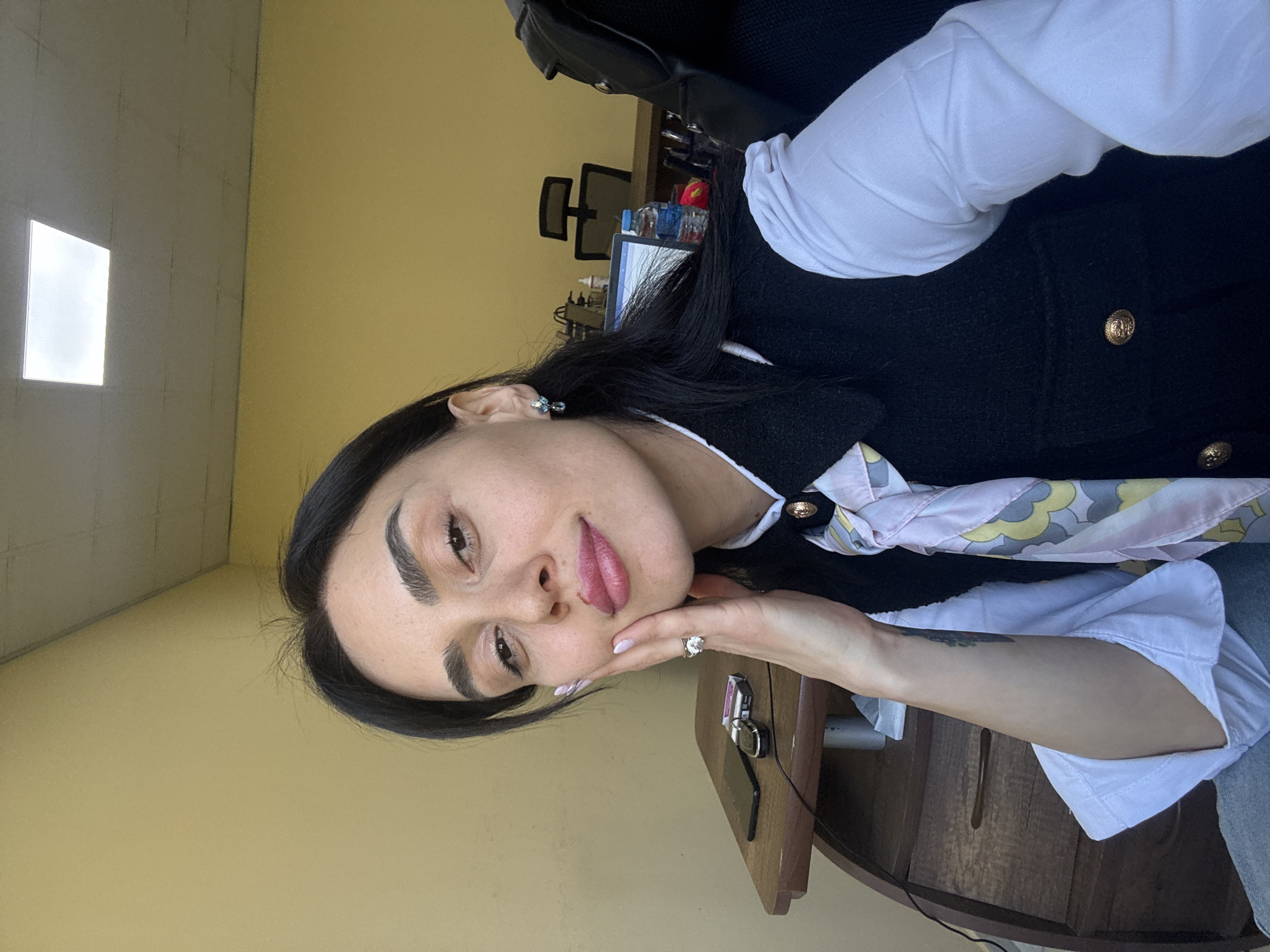 The image captures a young woman taking a selfie or posed portrait indoors, likely within an office environment in Tbilisi, Georgia. She has long dark hair, dark eyebrows, and is looking directly at the camera with a calm, subtle expression. Her lips are painted with a pink lipstick. She wears a white long-sleeved shirt underneath a dark-colored jacket or blazer featuring golden buttons. A light-colored scarf with a swirling pattern in shades of yellow, gray, and pink is tied around her neck. She accessorizes with small light-colored earrings and a prominent ring with a large clear stone on her left ring finger, her hand resting near her chin.
On the dark wooden desk in the foreground, a black smartwatch and keys are visible. The background reveals typical office elements: a black office chair, a computer monitor, and other items on a desk. The walls are a light yellow-beige color, and a bright rectangular light source or window is visible in the upper left portion of the image, suggesting a well-lit daytime setting. No visible text is present.