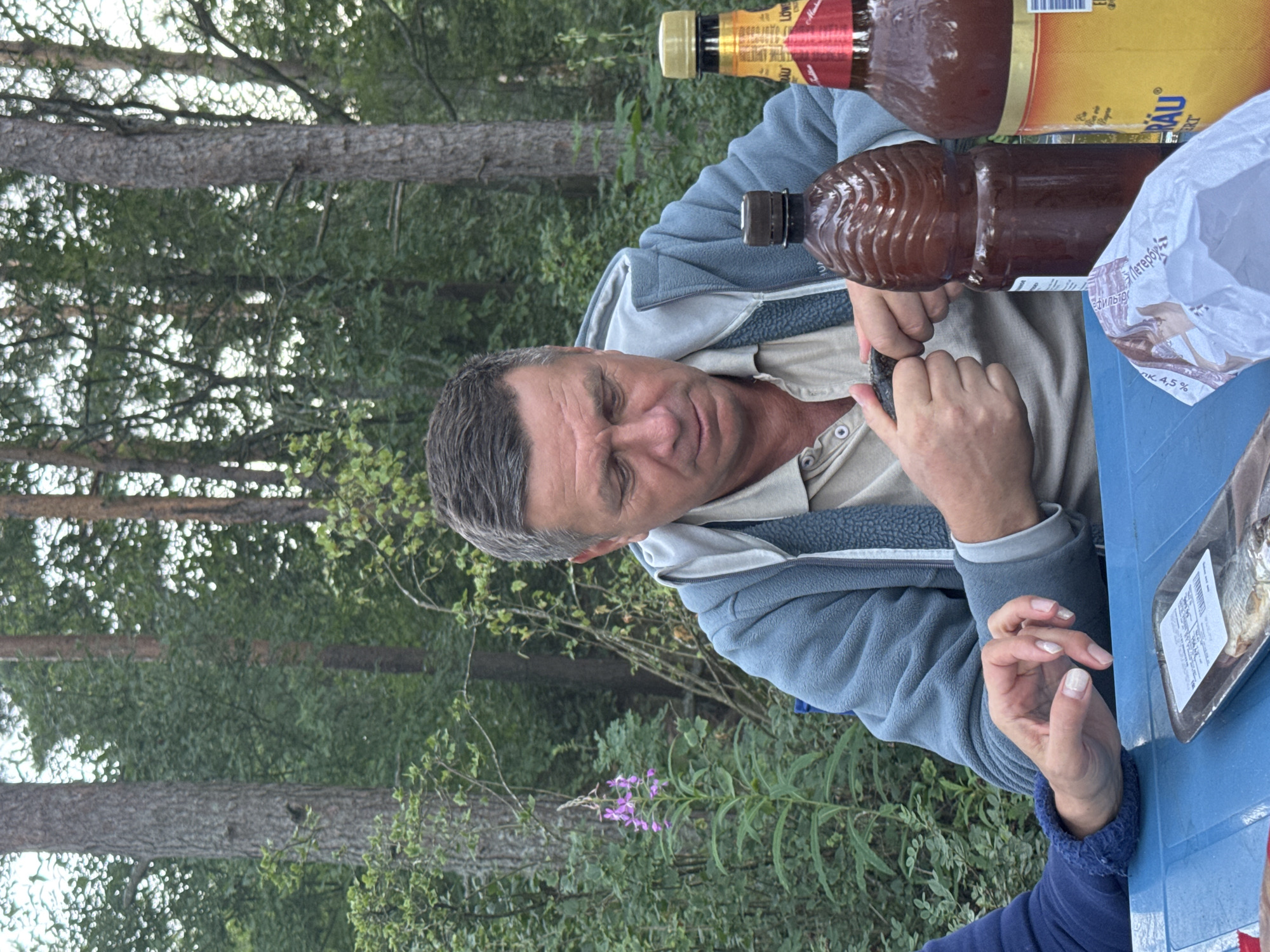 In Zelenaya Roshcha, Russia, a man is seated at a blue plastic table outdoors, appearing to be in a wooded or park-like area with tall trees and green foliage in the background. He is wearing a light-colored collared shirt and a blue hooded jacket. His gaze is directed downwards towards his hands, which are engaged in an action that is not clearly discernible but involves holding small objects. On the table in front of him are two brown plastic bottles, one with a ridged design, and a bottle of beer with a yellow label. A package of smoked fish in a plastic tray and a white plastic bag are also visible on the table. To the left of the man, a woman's arms and hands are visible, resting on the table. Her left arm is clad in a dark blue sleeve. The overall impression is that of a casual outdoor gathering.