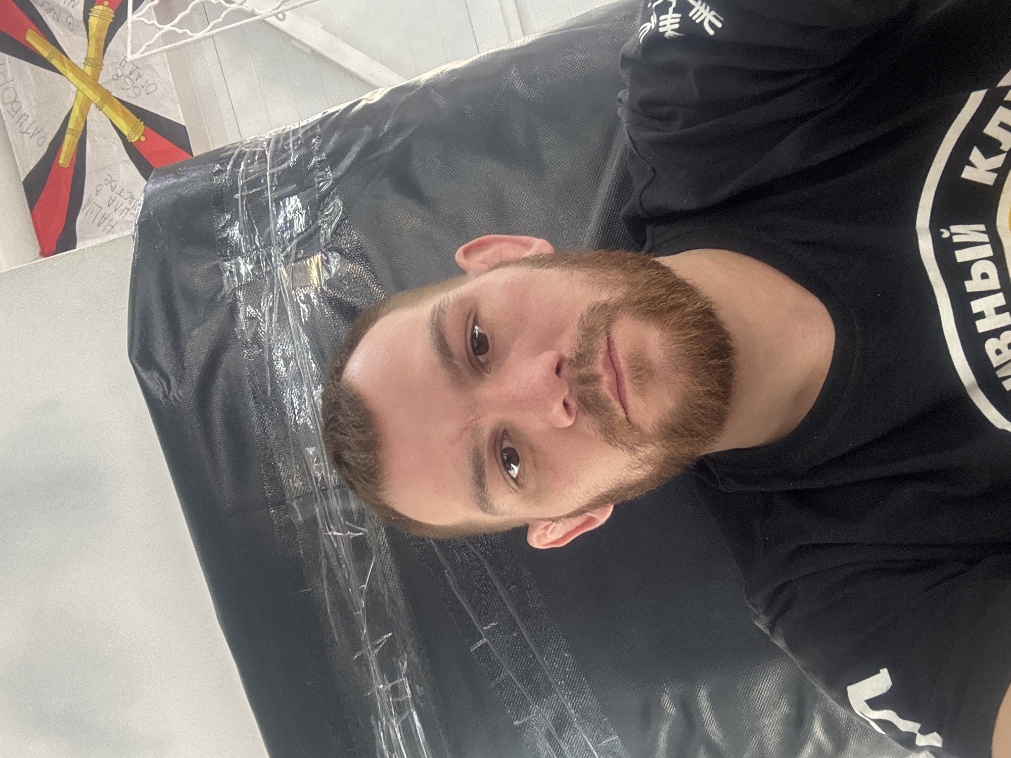 The image is a close-up, slightly angled selfie of a Caucasian man with a reddish-brown beard and short hair. He is wearing a black t-shirt with Cyrillic text and a logo in white. The background includes a black, tarp-like material covered in clear plastic wrap. A flag with a crossed-cannon design and Cyrillic inscriptions is visible in the upper left corner. The location is likely Perm, Russia, based on the flag's design and the Cyrillic text on the t-shirt.